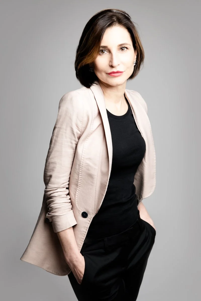 Una donna con capelli castani corti e occhi chiari, indossa una giacca beige, una maglietta nera e pantaloni neri, posa con le mani nelle tasche, sfondo grigio chiaro.