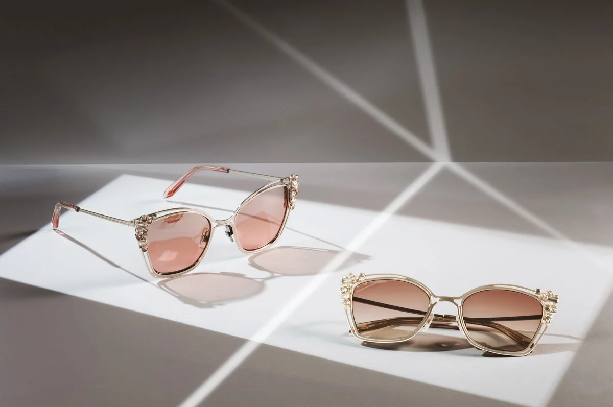 Due paia di occhiali da sole con montature decorate, uno con montatura rosa e l'altro con montatura dorata, posizionati su una superficie bianca con una luce che crea ombre