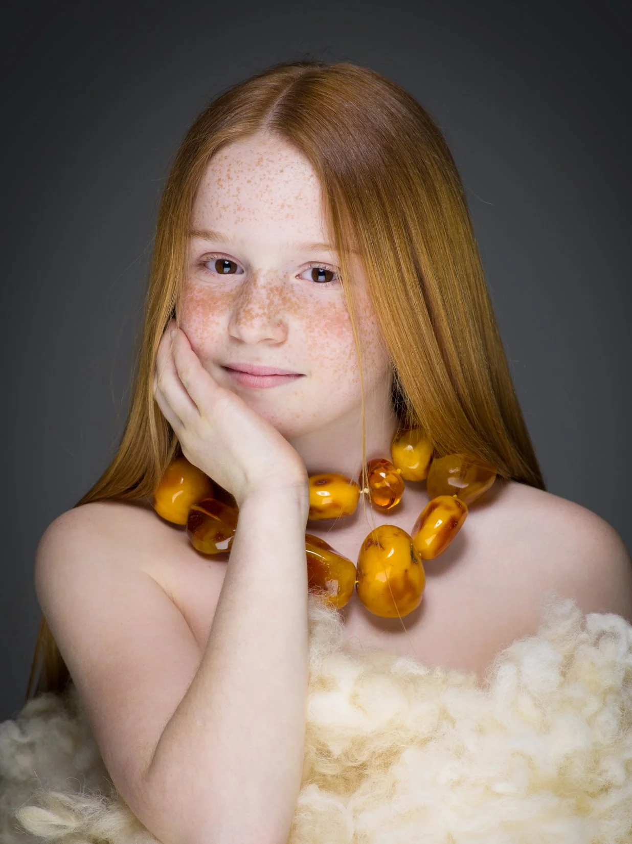 Ragazza con capelli rossi, pelle ricoperta di lentiggini, indossa una collana di grandi pietre ambra e un abito di pelliccia beige, in posa con la mano sul viso, sfondo grigio scuro.
