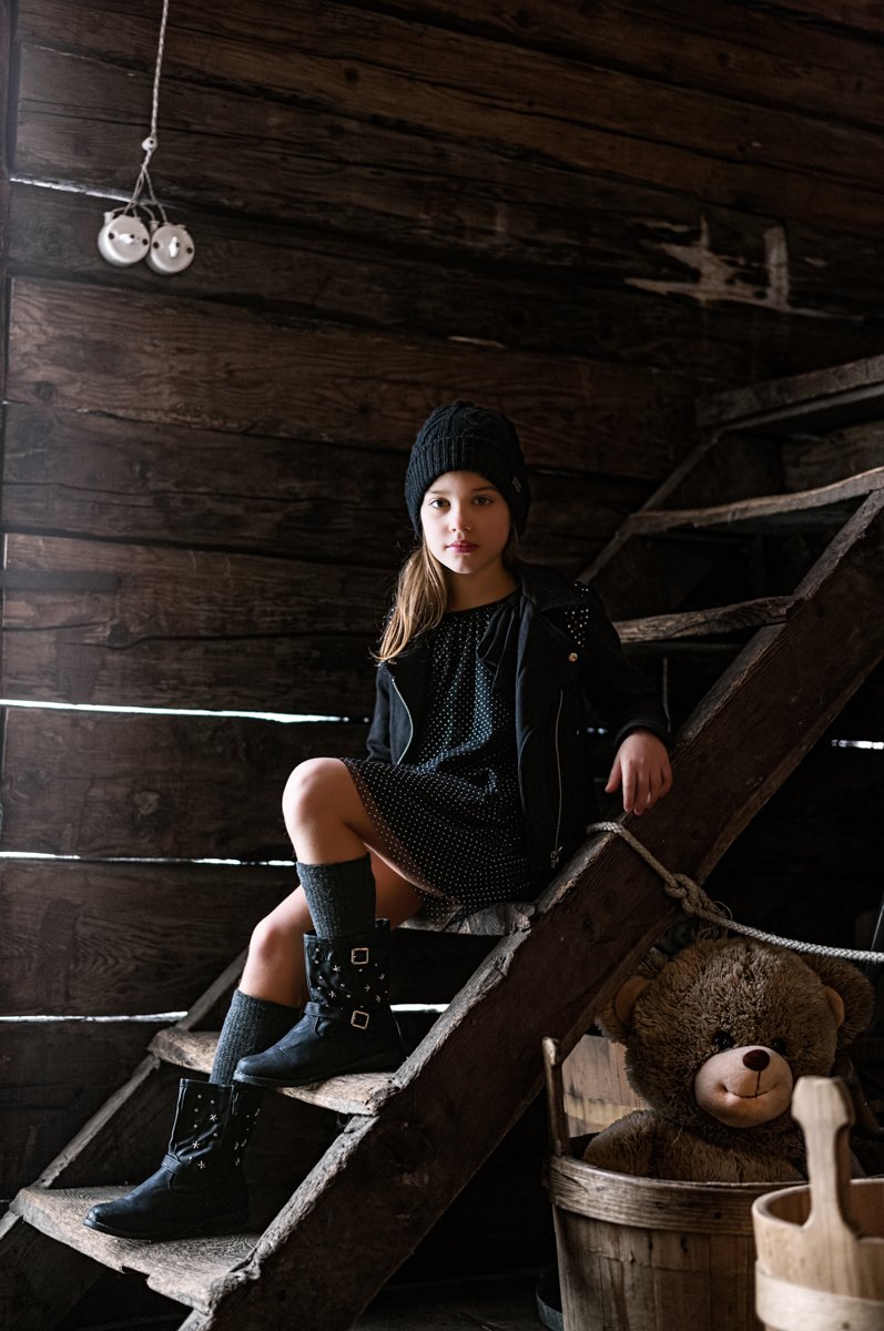Una giovane ragazza seduta su una scala in legno in un ambiente rustico, indossa un cappello nero, vestito scuro con maniche a tre quarti, giacca nera e scarpe con borchie. Dietro di lei, una parete in legno scuro e una lampada a due globi appesa al 