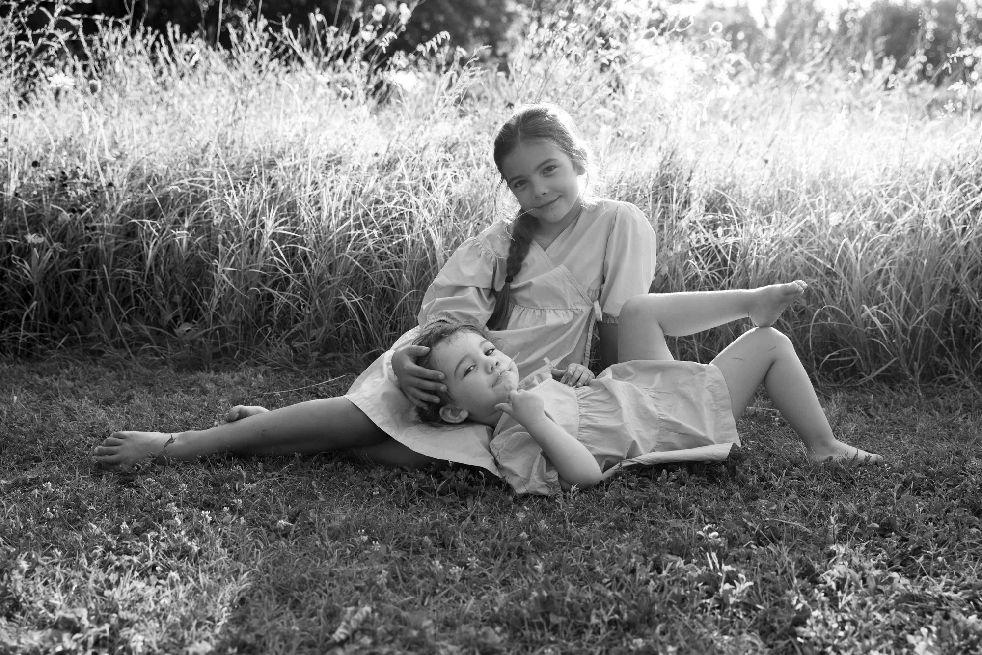 Due bambine che giacciono sull'erba in un campo, uno sdraiata con il braccio sotto la testa e l'altro che si sostiene con i gomiti, in uno scenario naturale e soleggiato.