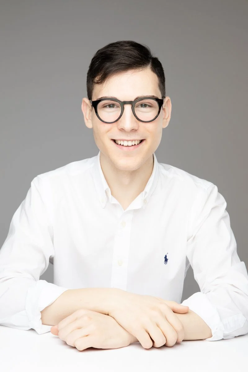 Giovane uomo con occhiali neri, camicia bianca e sorriso, seduto davanti a uno sfondo grigio.