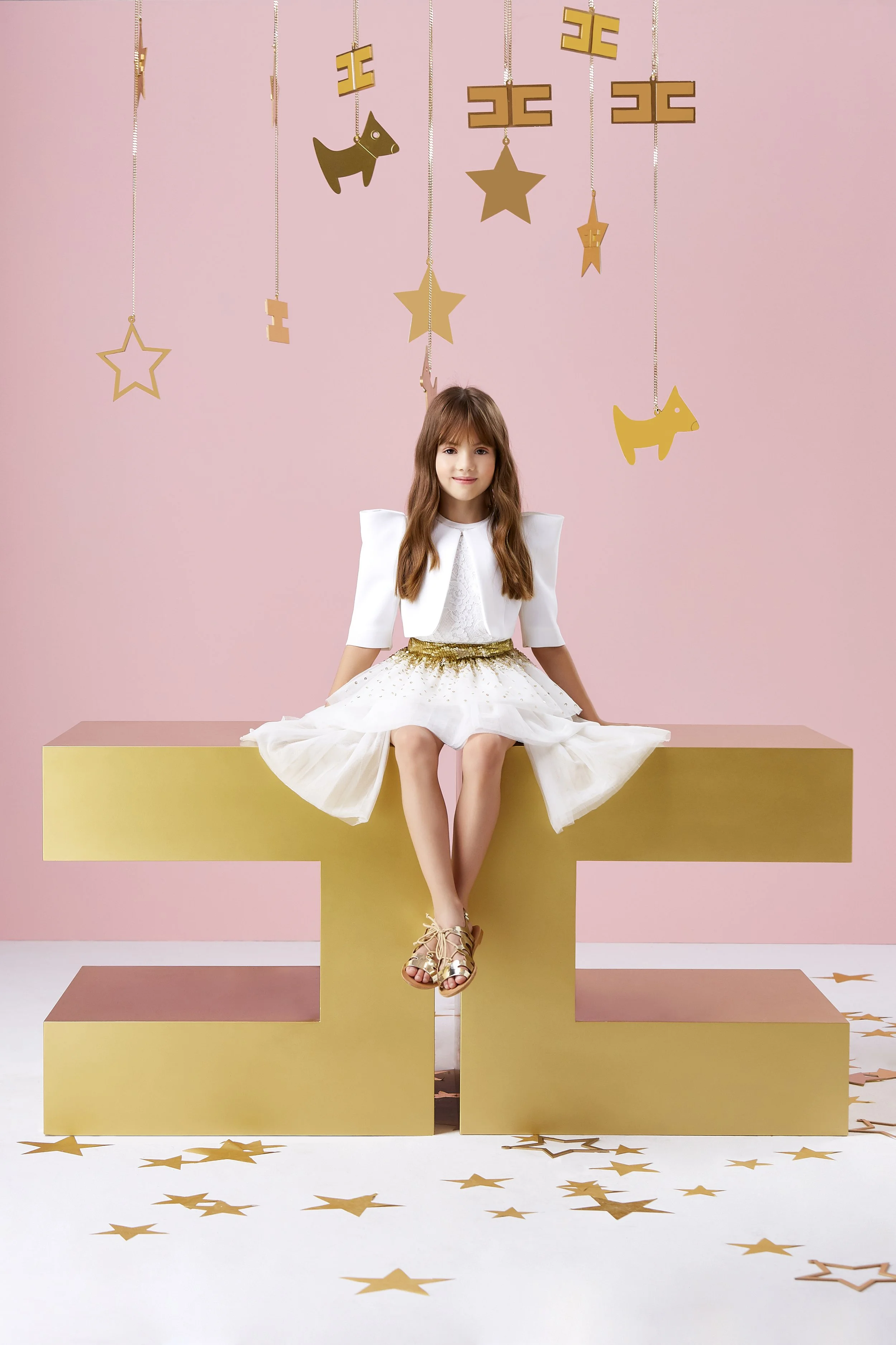 Una ragazzina seduta su una grande lettera 'T' color oro, con un cappello a forma di stella dorata, un maglione bianco e una gonna di tulle bianca con dettagli dorati. Sullo sfondo, pareti rosa e decorazioni in metallo oro a forma di stelle, stelle e