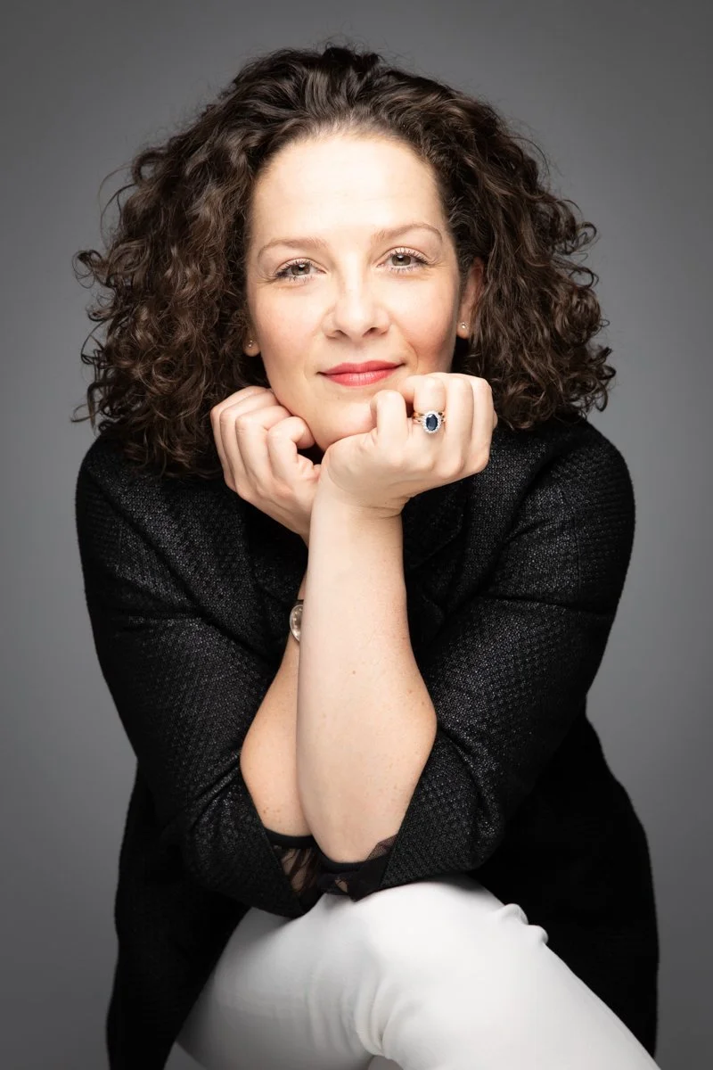 Ritratto di una donna con capelli riccioli marroni, che sorride leggermente, appoggiando il viso alle mani chiuse a pugno. Indossa un blazer nero e ha un anello con gemma blu.