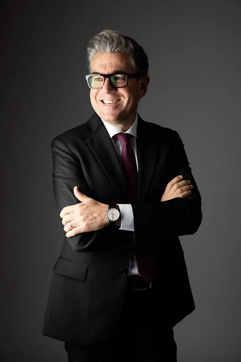 Uomo d'affari sorridente con occhiali, vestito elegante, braccia incrociate, sfondo grigio.
