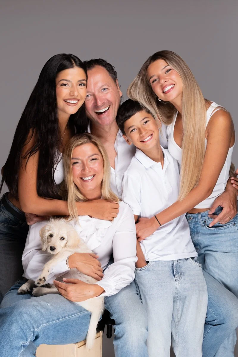 Una famiglia sorridente composta da due ragazze, due ragazzi, un uomo e una donna, con un cane bianco, tutti vestiti in bianco e jeans, posano felici contro uno sfondo grigio.