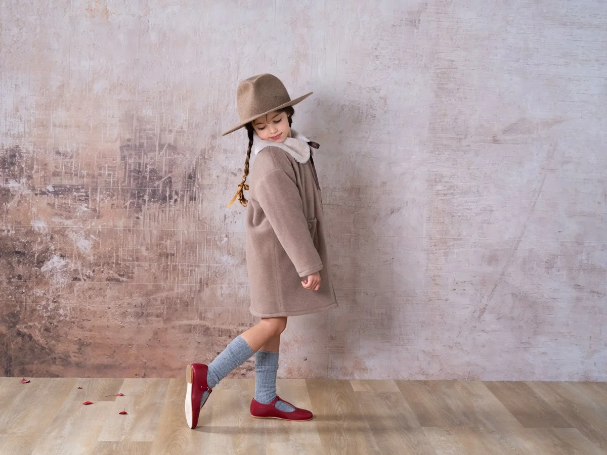 Bambina con cappello, cappotto beige, calzoni lunghi grigi, scarpe rosse e calzettoni grigi, che si ferma con una gamba sollevata contro uno sfondo di parete beige.