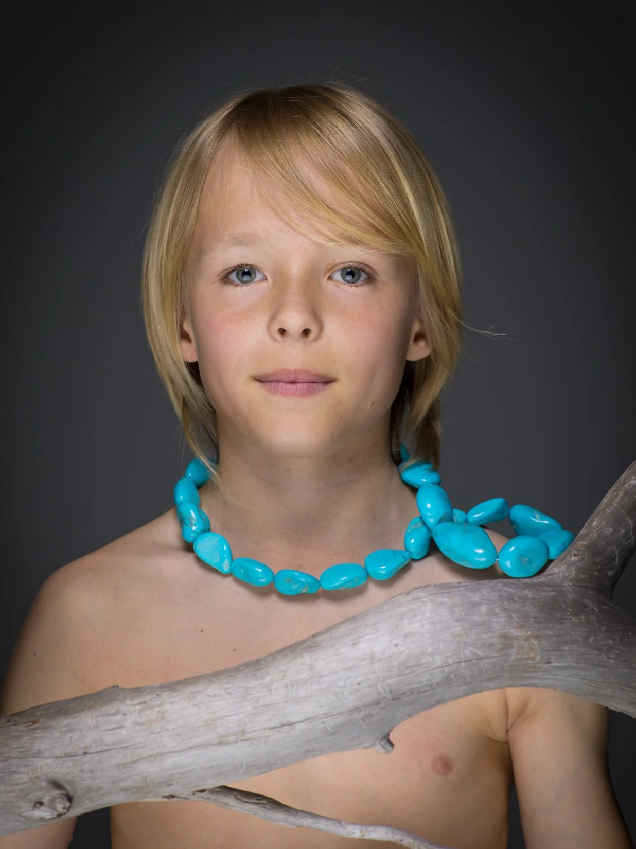 Ragazzo con capelli biondi, occhi azzurri, che indossa una collana di pietre turchesi, posizionato davanti a uno sfondo scuro. Ha una pelle chiara e un'espressione tranquilla.
