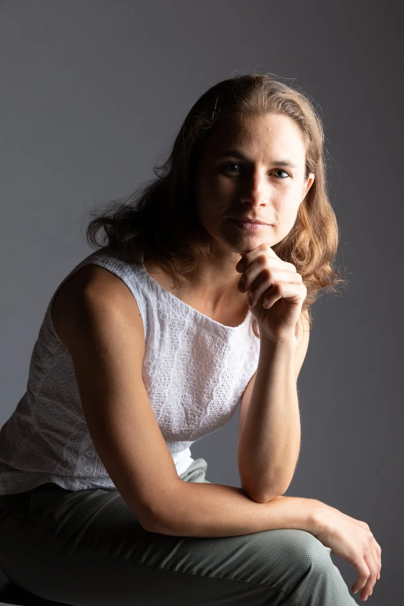 Ritratto di una donna con capelli castani chiari, abbigliata con una blusa bianca, seduta contro uno sfondo grigio, poggiando il volto sulla mano, con espressione serena.
