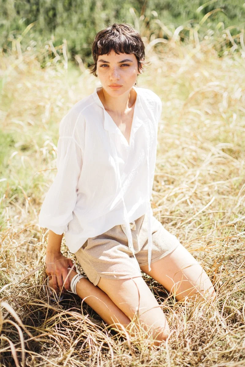 Una giovane donna con capelli corti e scuri, indossa una camicia bianca e pantaloncini beige, si trova in posizione inginocchiata in un campo di erba secca, sotto la luce del sole.