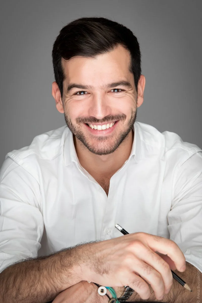 Uomo sorridente con camicia bianca, seduto con braccia appoggiate sul tavolo, tiene una penna in mano, sfondo grigio, immagine in primo piano.