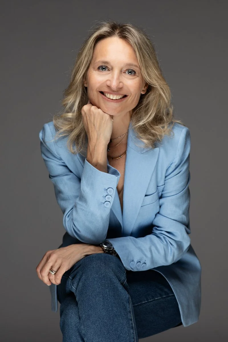 Donna sorridente con capelli biondi, indossa una giacca azzurra e jeans, mentre si siede con il braccio appoggiato sulla ginocchio e il mento sulla mano, sullo sfondo grigio.