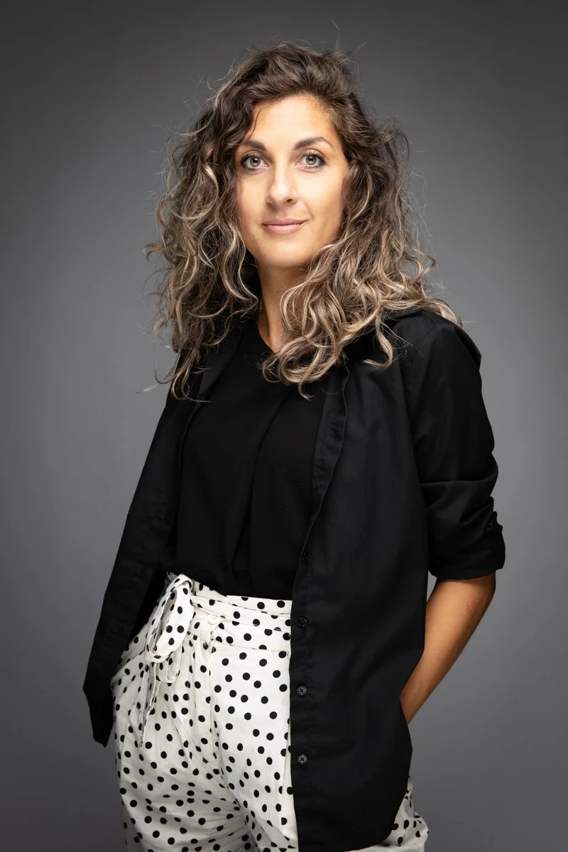Una donna con capelli ricci, abbigliata con una camicia nera e pantaloni banta con punti neri, posa su uno sfondo grigio