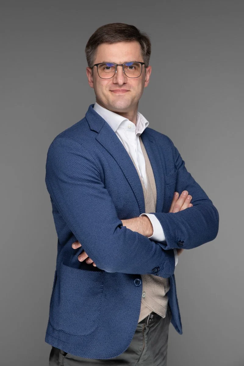 Uomo sorridente con occhiali e capelli castani, indossa una giacca blu, una camicia bianca e un maglione beige, posa con le braccia incrociate davanti a uno sfondo grigio.
