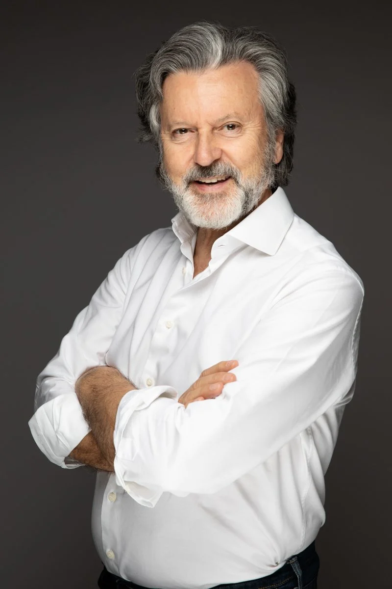 Uomo anziano con capelli grigi e barba, indossa una camicia bianca, sorride e ha le braccia incrociate contro uno sfondo grigio.