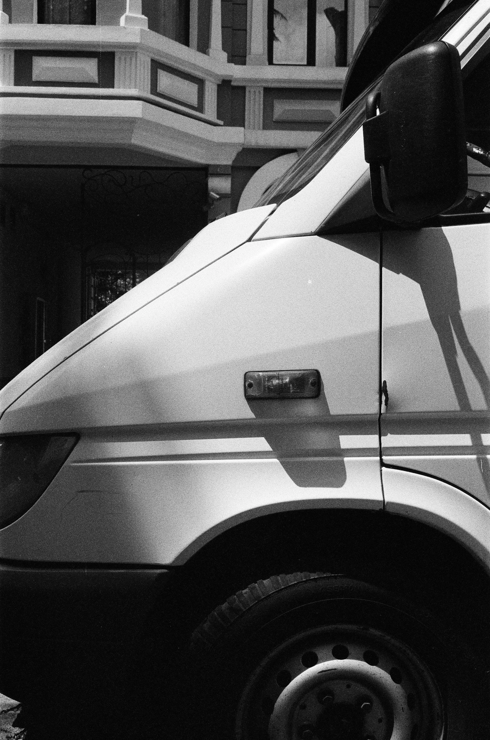 Black and white photo of the front part of a commercial van or truck, showing the side mirror, a headlight, and part of the wheel, with a building and trees in the background.