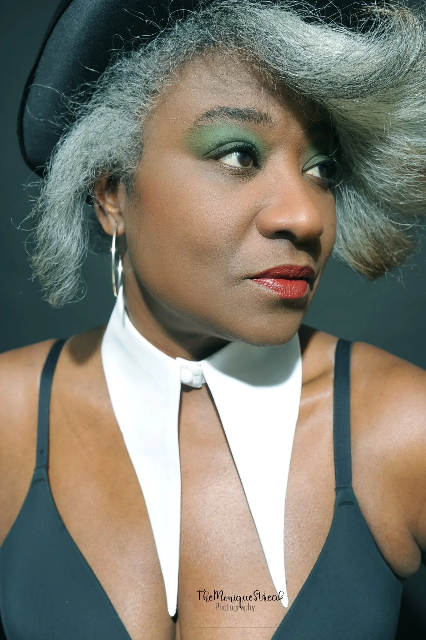 Close-up of a woman with gray and black hair, wearing a black top with thin straps and a white neck accessory, posing against a dark background.