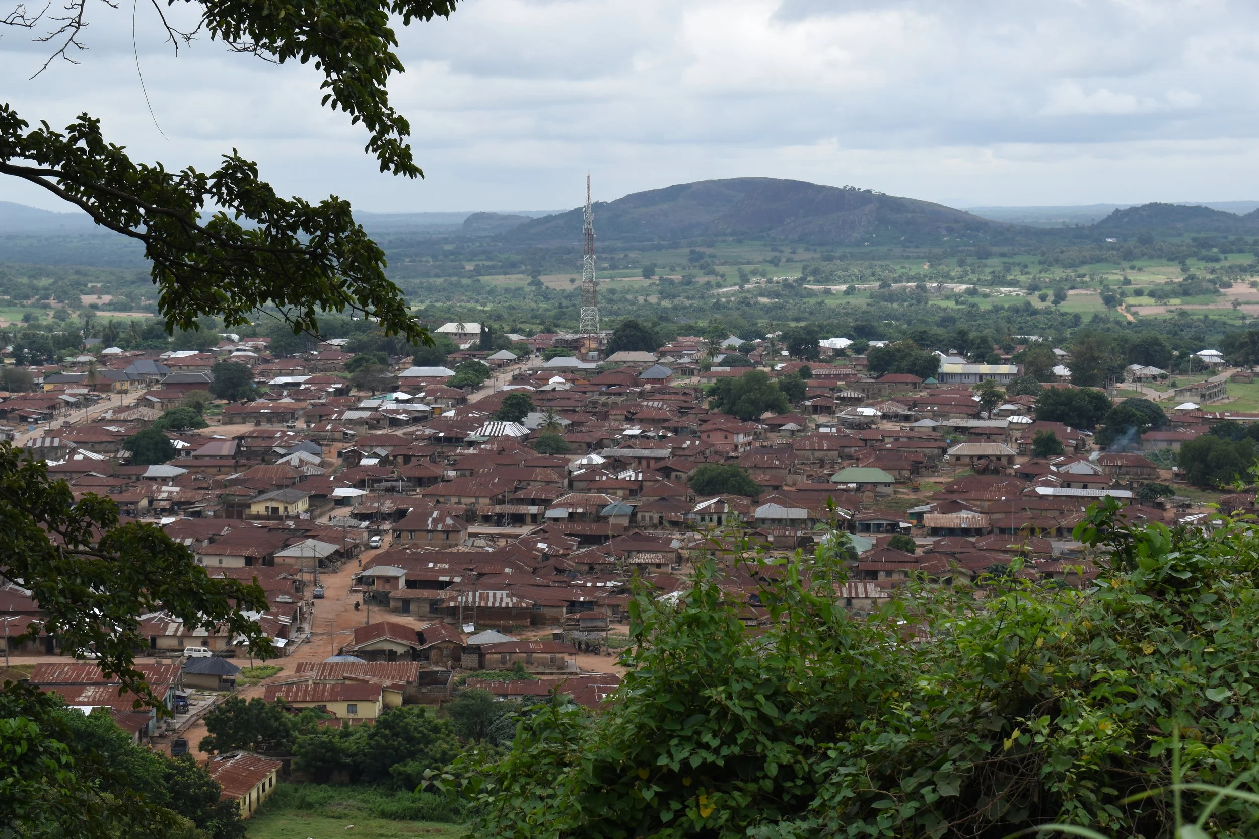 A view of a small town with numerous houses and buildings with rust-colored roofs, nestled among green trees and shrubs, with a tall communication tower in the background. Behind the town, there is a large hill or mountain, and the sky is mostly cloudy.