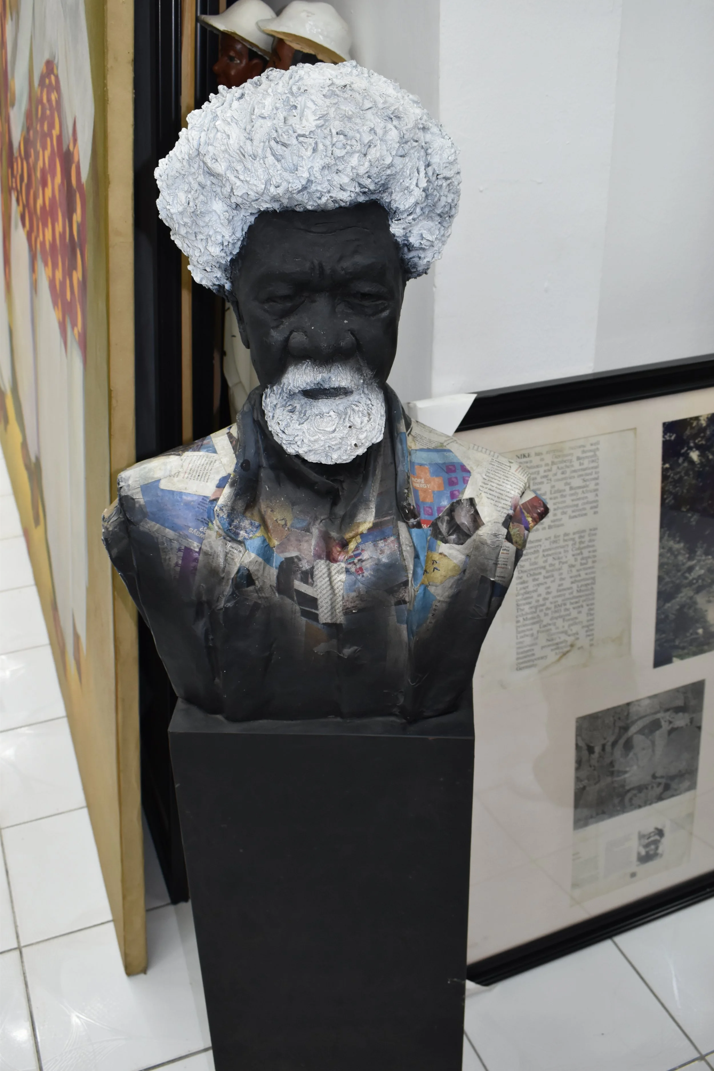 Bust of a man with dark skin, white hair, and a white beard, wearing a newspaper shirt, with additional sculptures and images in the background.