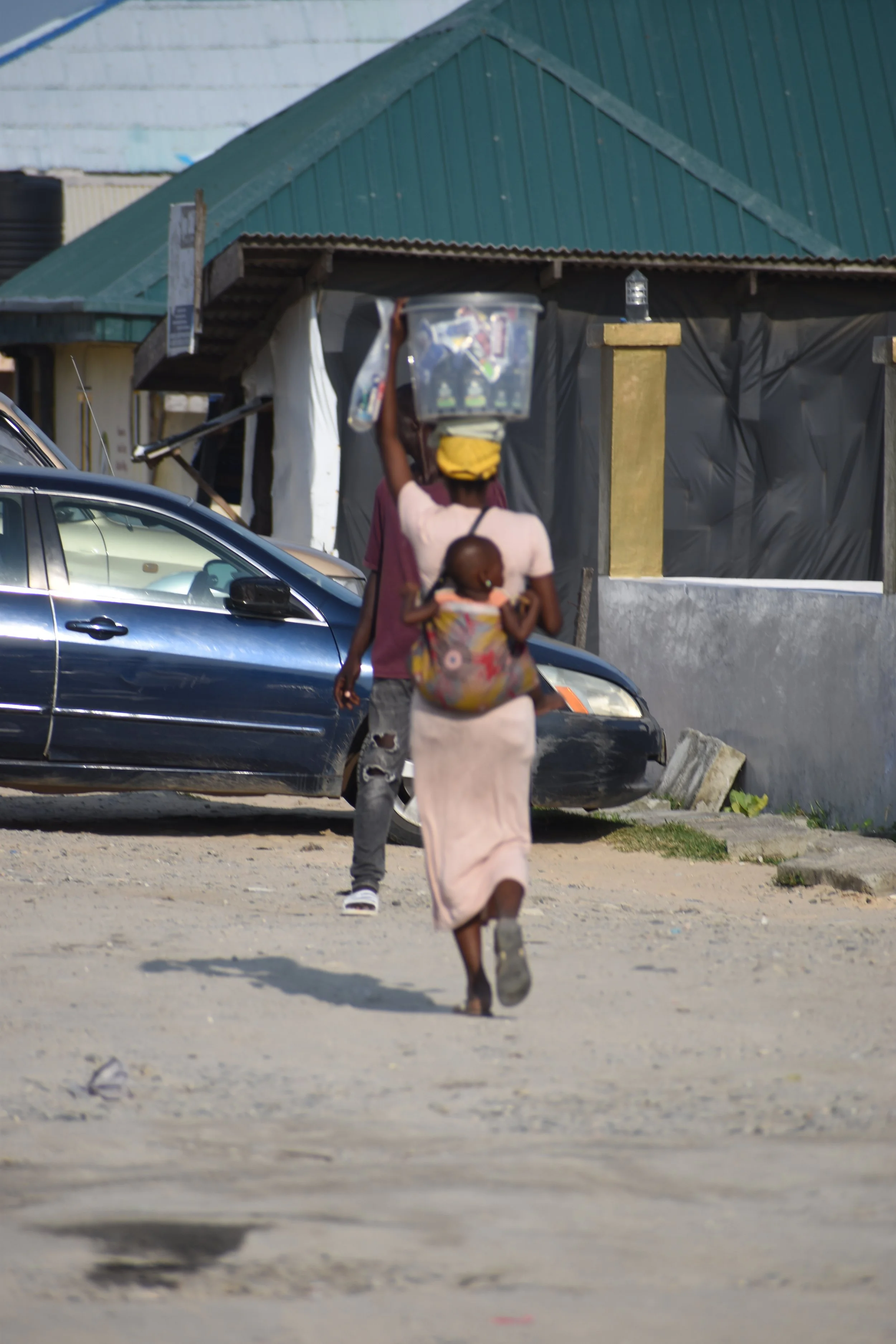 A woman carrying a child on her back and balancing a plastic container on her head while walking on a dirt road in front of a blue and green building with parked cars nearby.