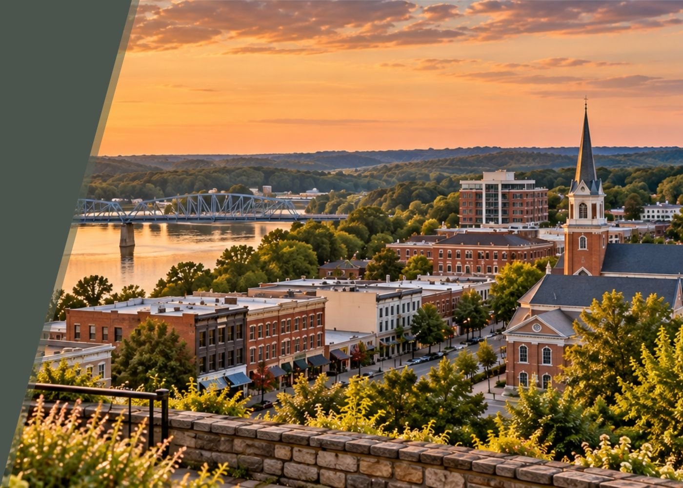 Sunset over a small town with a river, bridge, historic brick buildings, a church with a steeple, and tree-lined streets