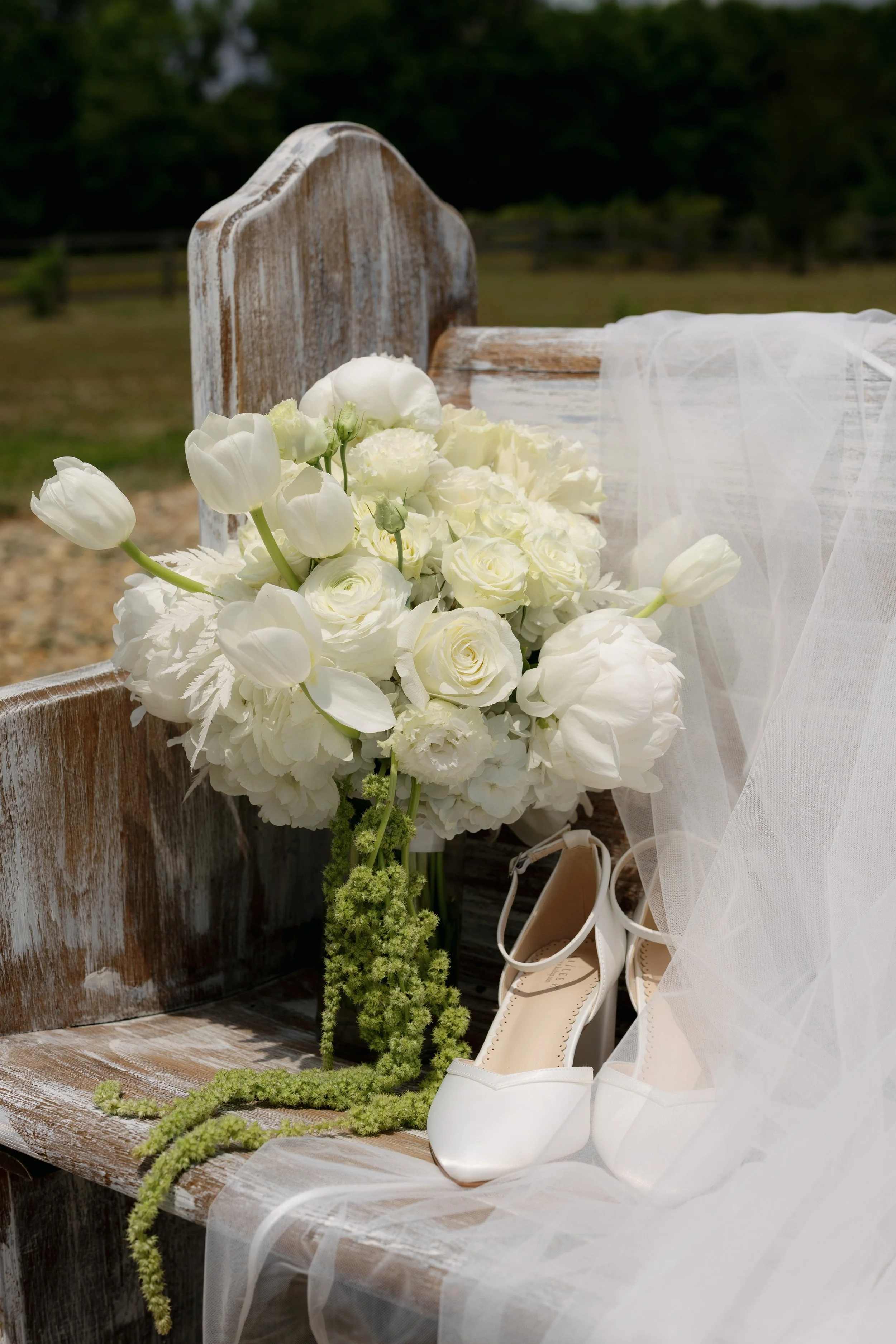 White bridal bouquet beside a rustic wooden bench, white wedding shoes, and flowing veil outdoors