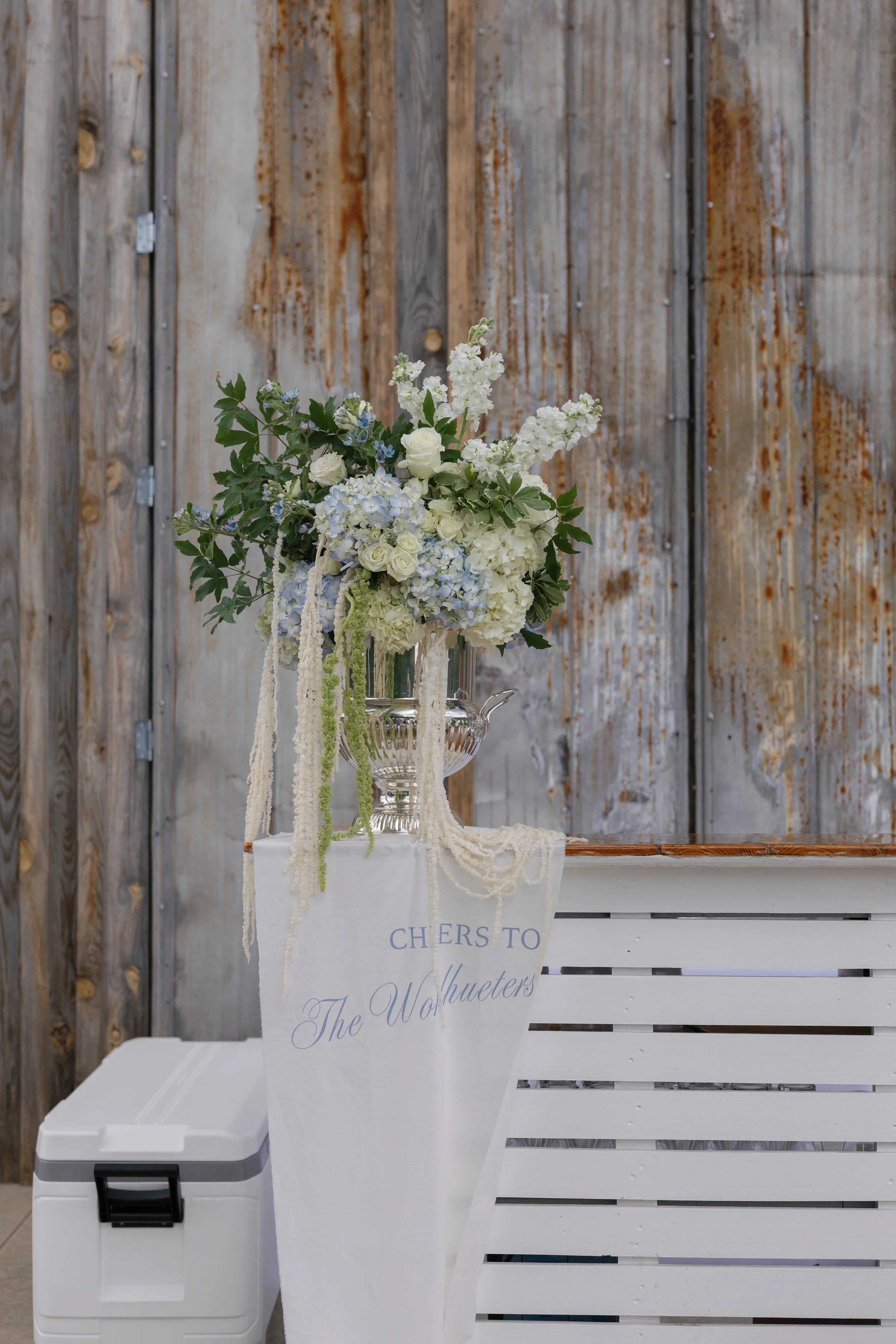 Elegant white and pale blue floral arrangement in a silver vase on a rustic wedding dessert table.