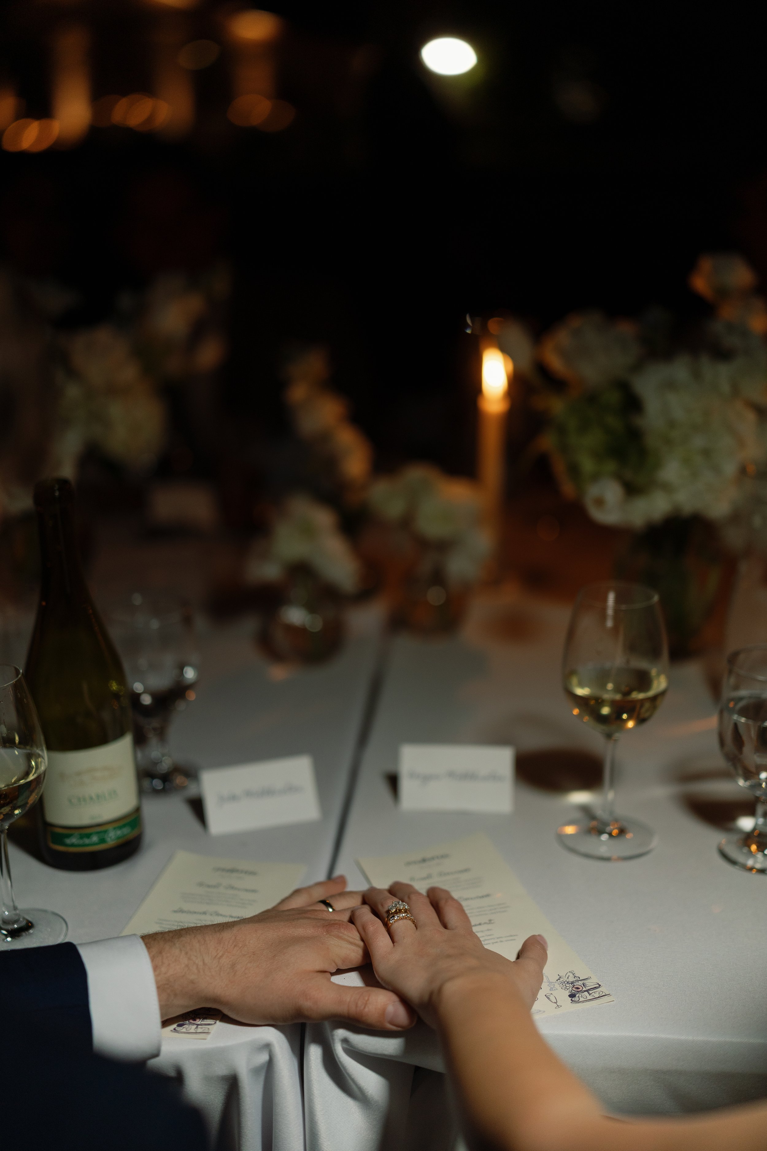 Close-up of a couple holding hands at a candlelit wedding reception table with wine bottles, glasses, and menus.