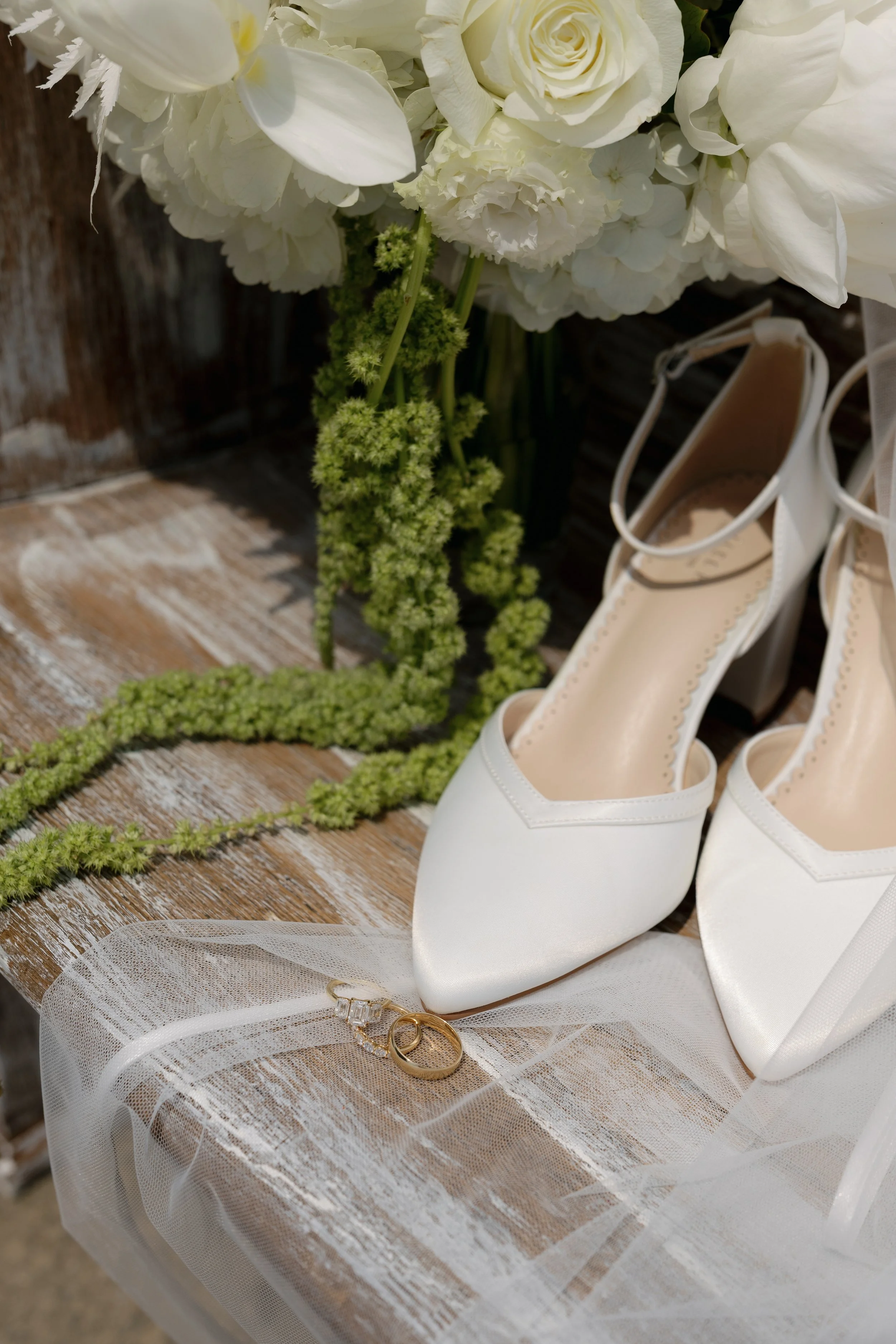 White bridal shoes, wedding rings, and white floral bouquet arranged on rustic wood with lace fabric.