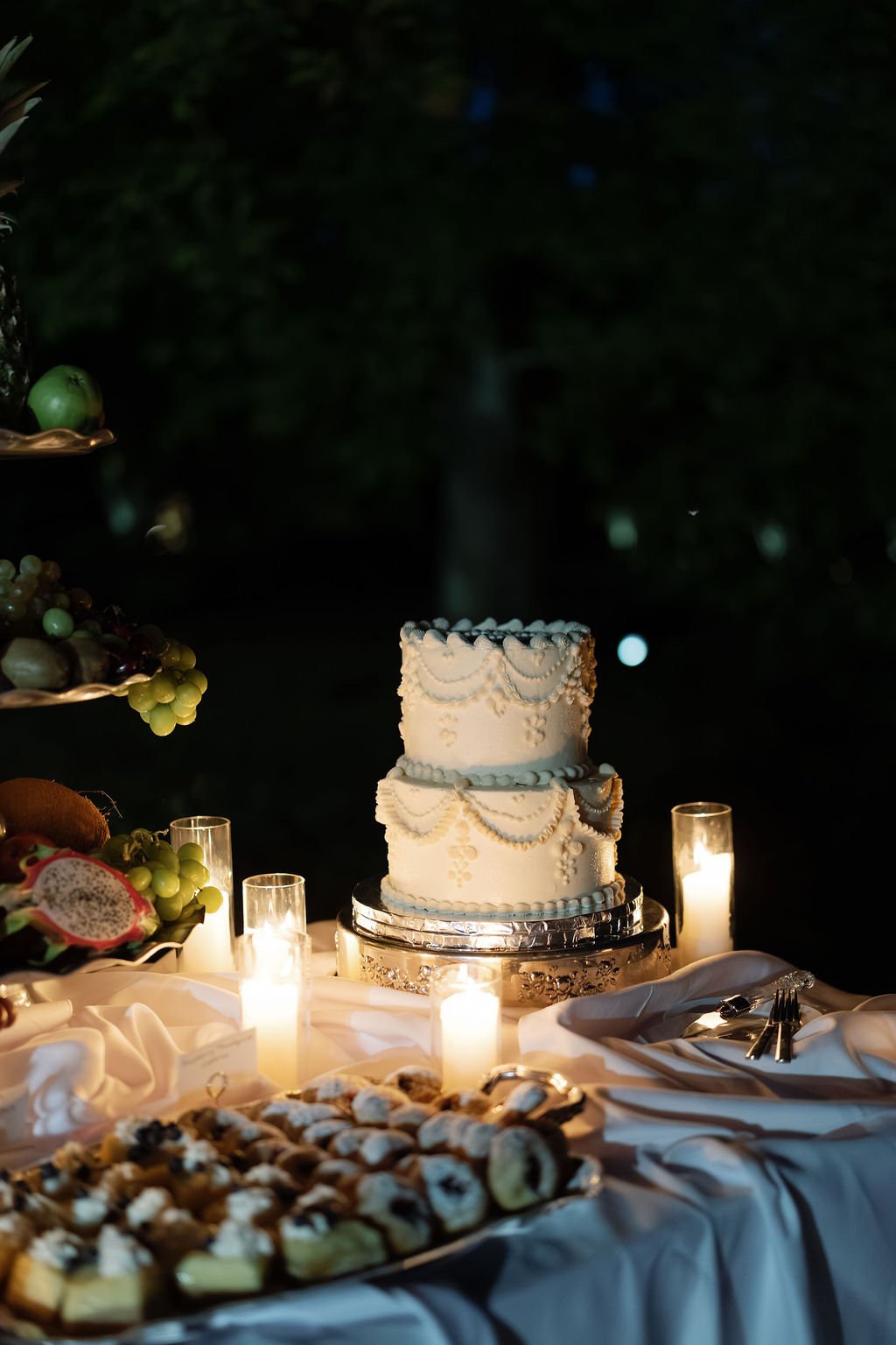 Elegant three-tier wedding cake with piped icing on a candlelit reception table surrounded by fruit and pastries