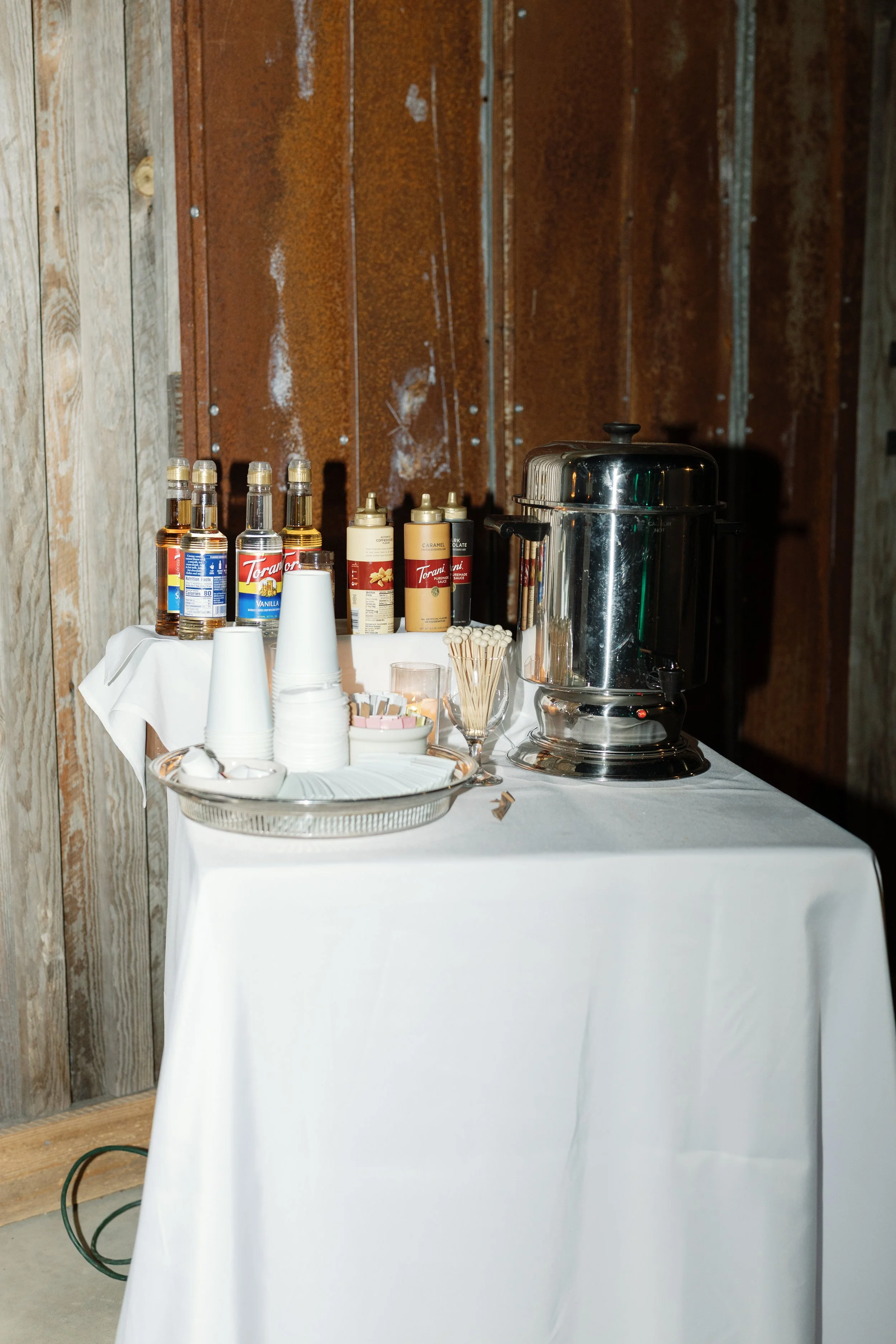Coffee and hot chocolate station with flavored Torani syrups, creamers, cups, toppings, and a stainless steel beverage dispenser.