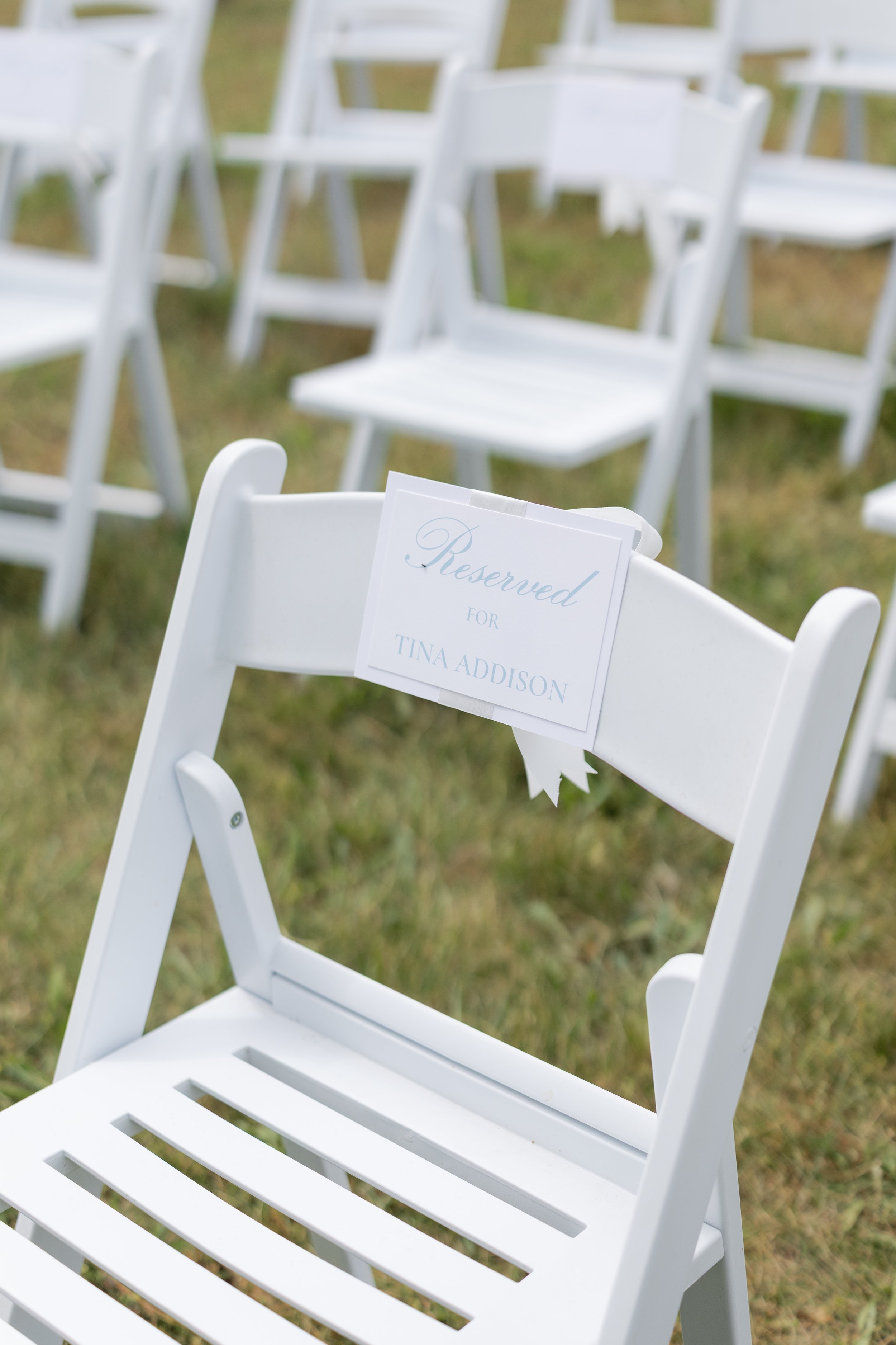 White outdoor ceremony chairs on grass, including a reserved sign for Tina Addison.