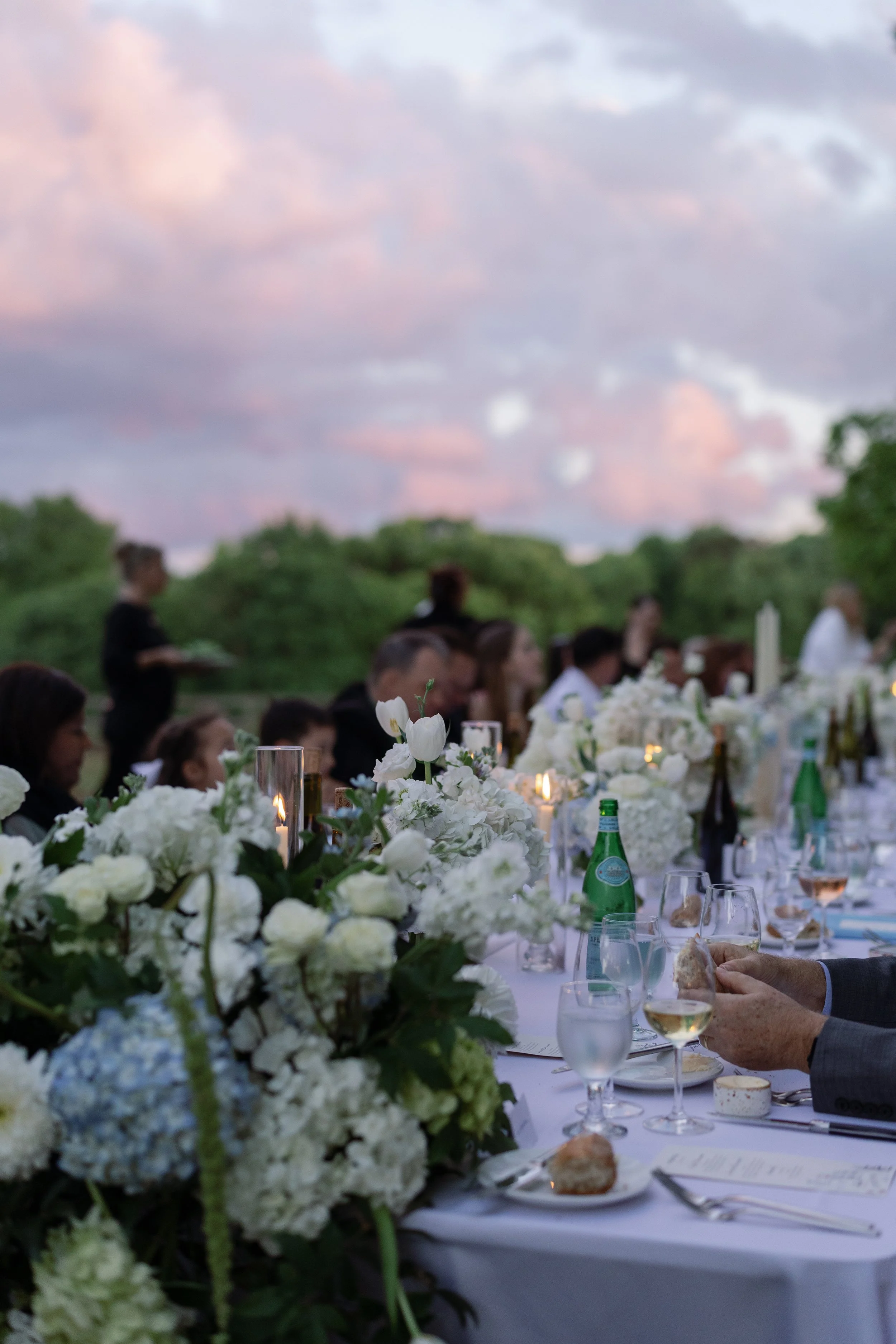 Outdoor dinner party with white floral centerpieces, candles, wine glasses, and guests beneath a pastel evening sky