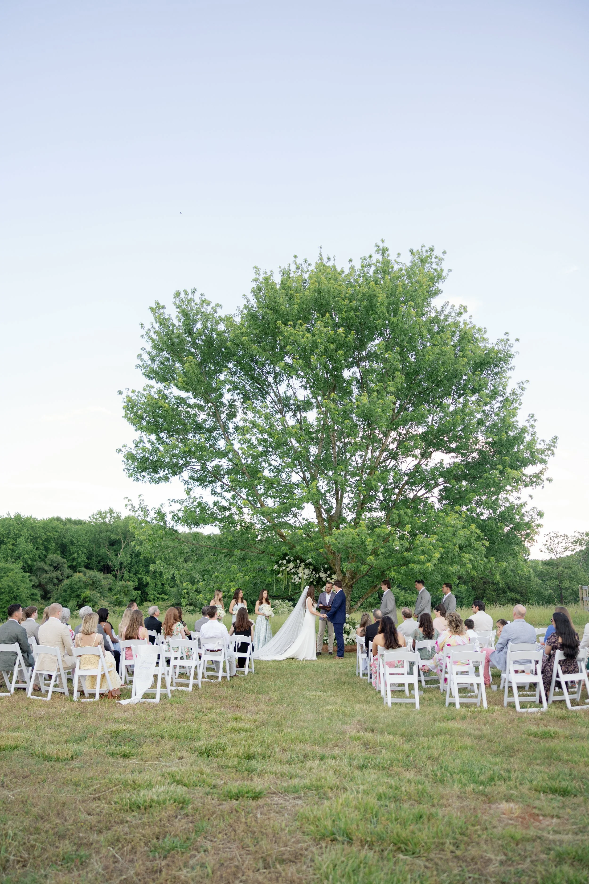 Outdoor wedding ceremony under a large tree, with the bride and groom at the altar and guests seated in white chairs.