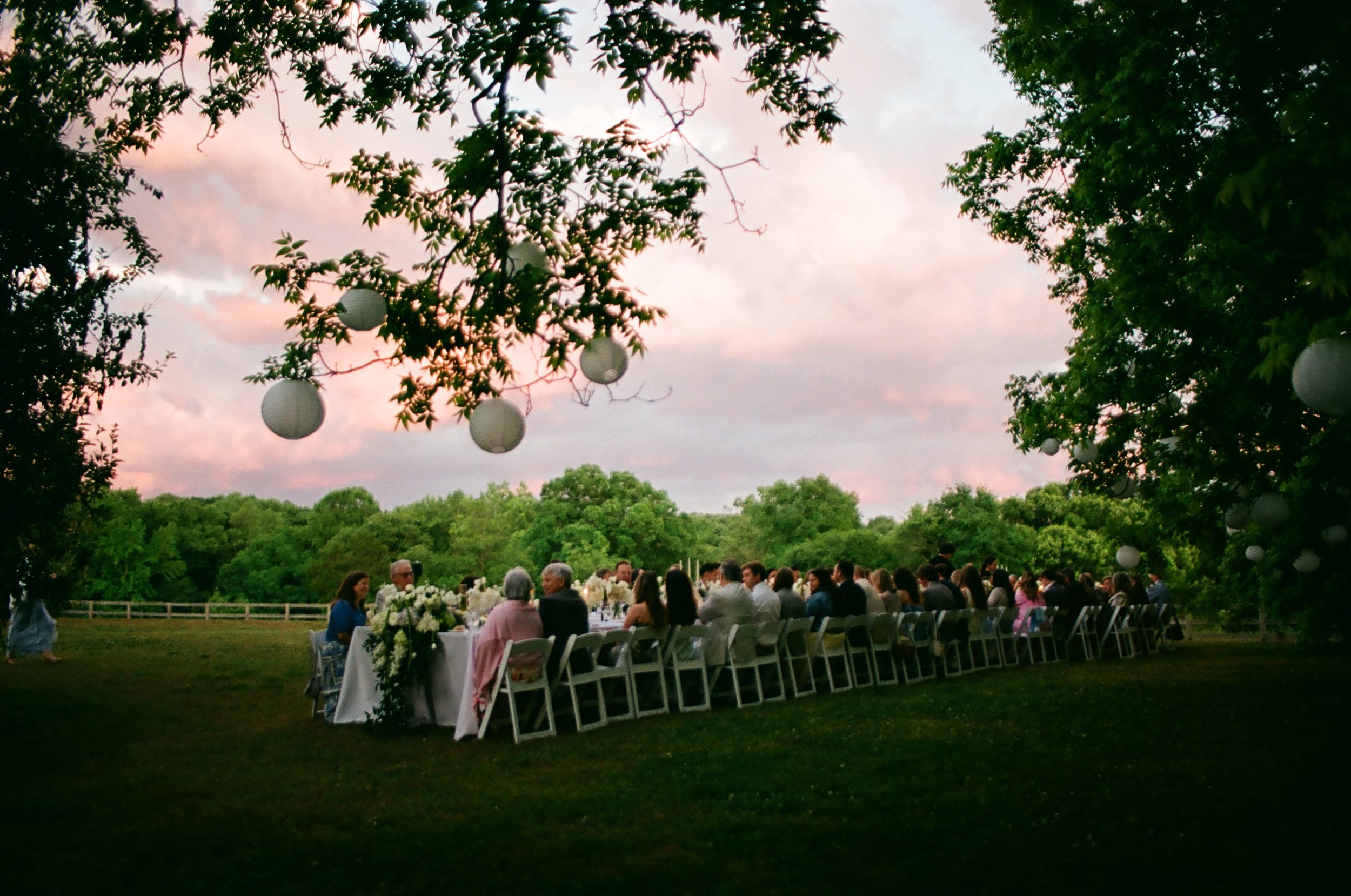 Outdoor wedding reception with guests seated at decorated tables beneath trees and white lanterns at sunset