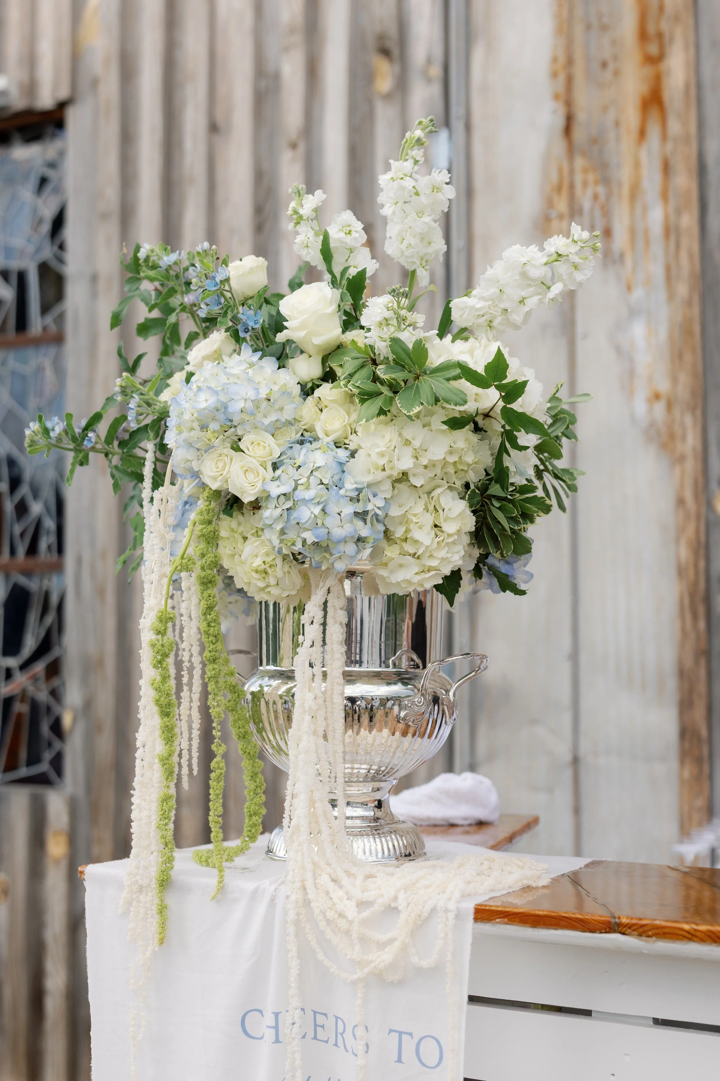 Elegant white and pale blue floral arrangement with roses, hydrangeas, and cascading flowers in a silver urn on a draped table