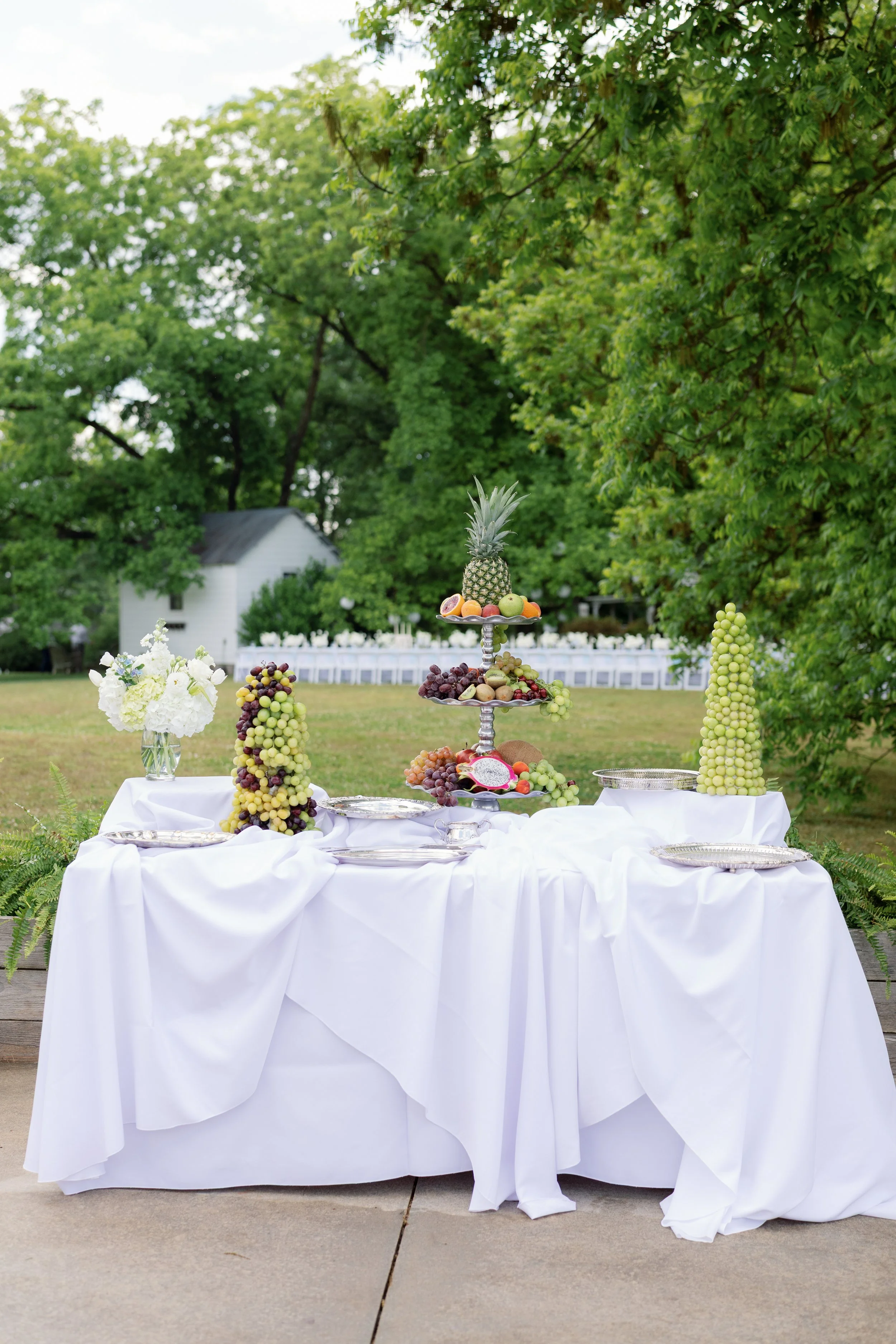 Outdoor event dessert table with white linens, fruit displays, pineapple centerpiece, and floral arrangement in a garden