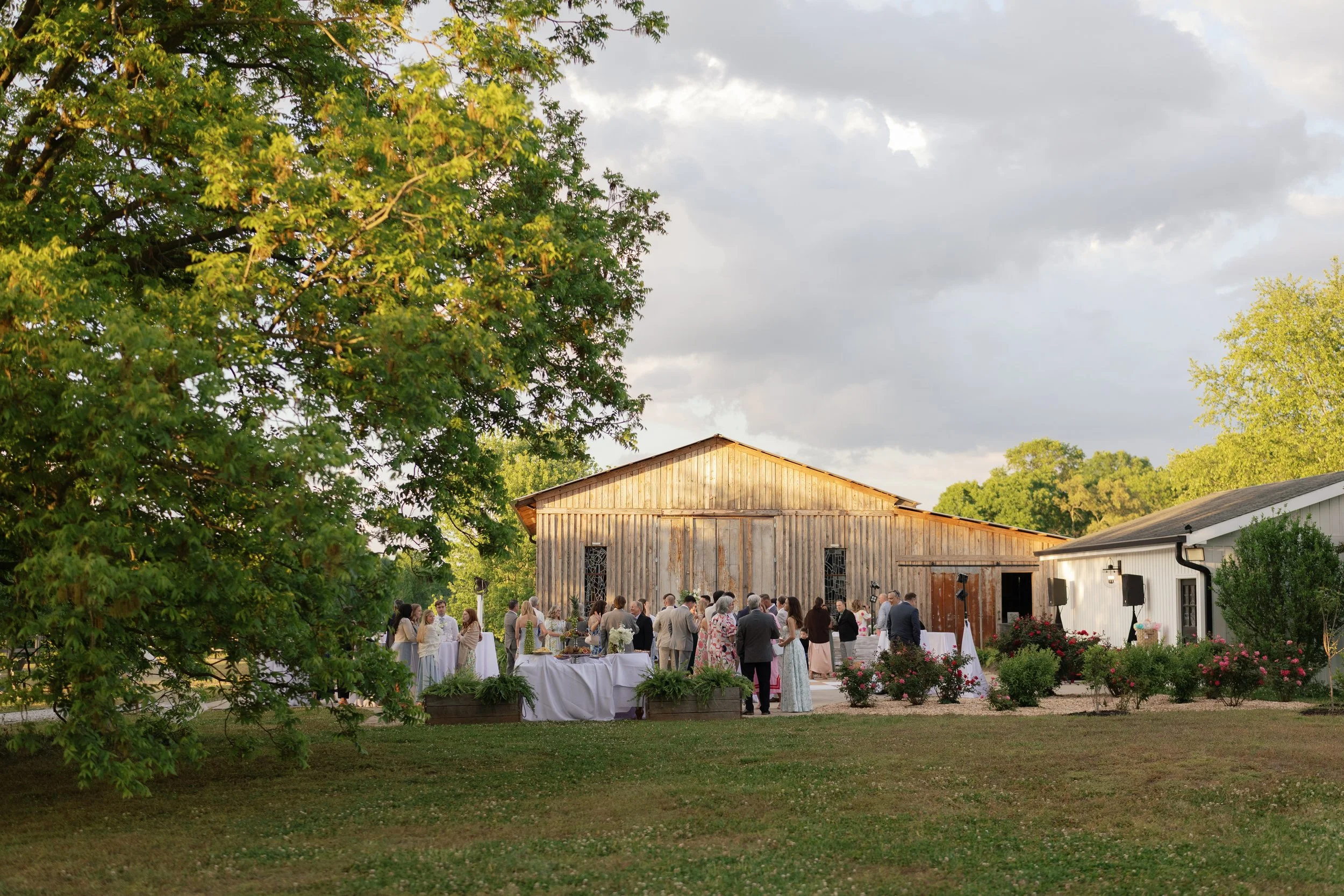 Outdoor wedding reception with guests, buffet tables, and floral arrangements beside a rustic wooden barn.