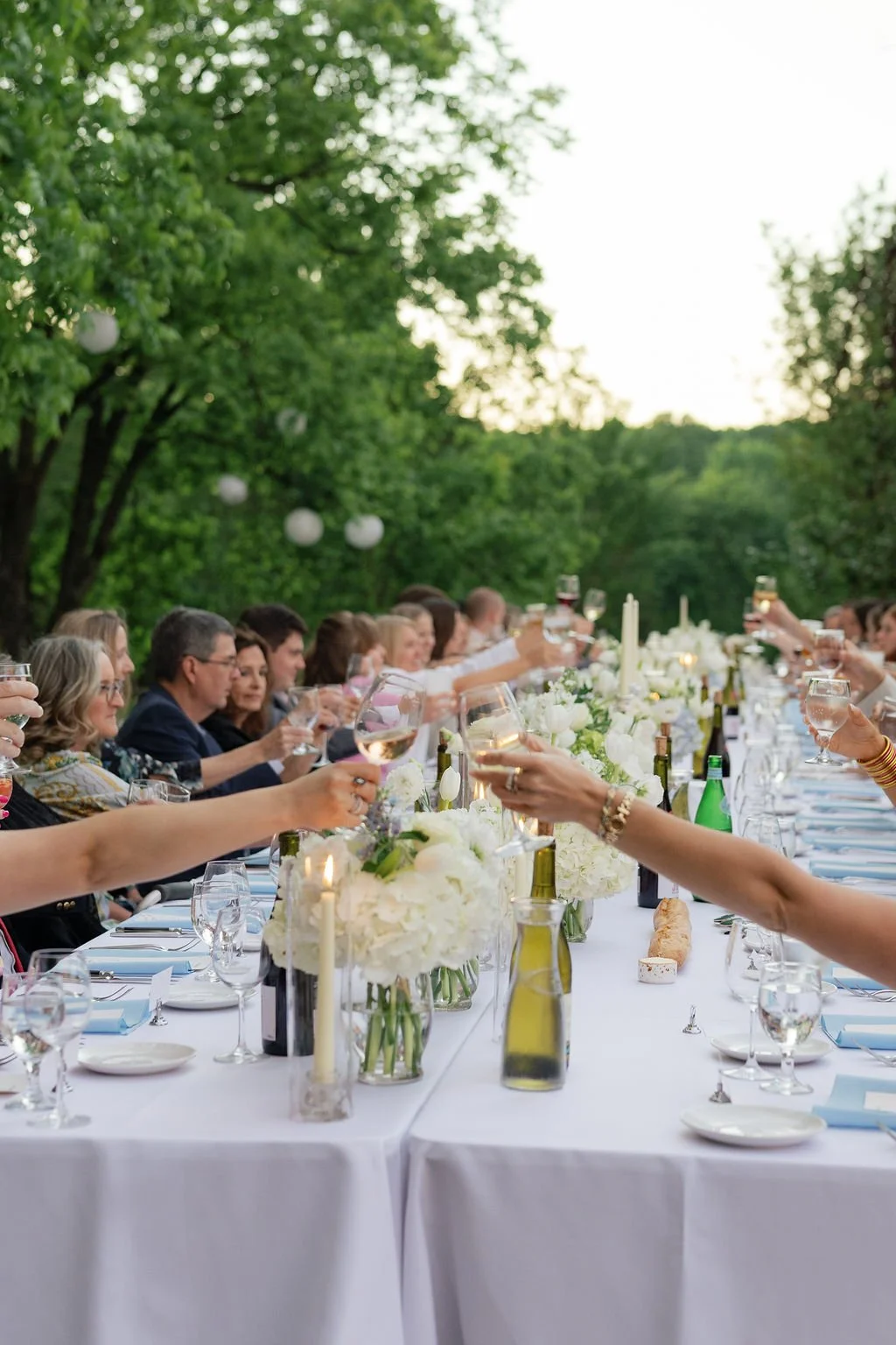 Guests raise wine glasses at an elegant outdoor dinner table decorated with white flowers and candles.