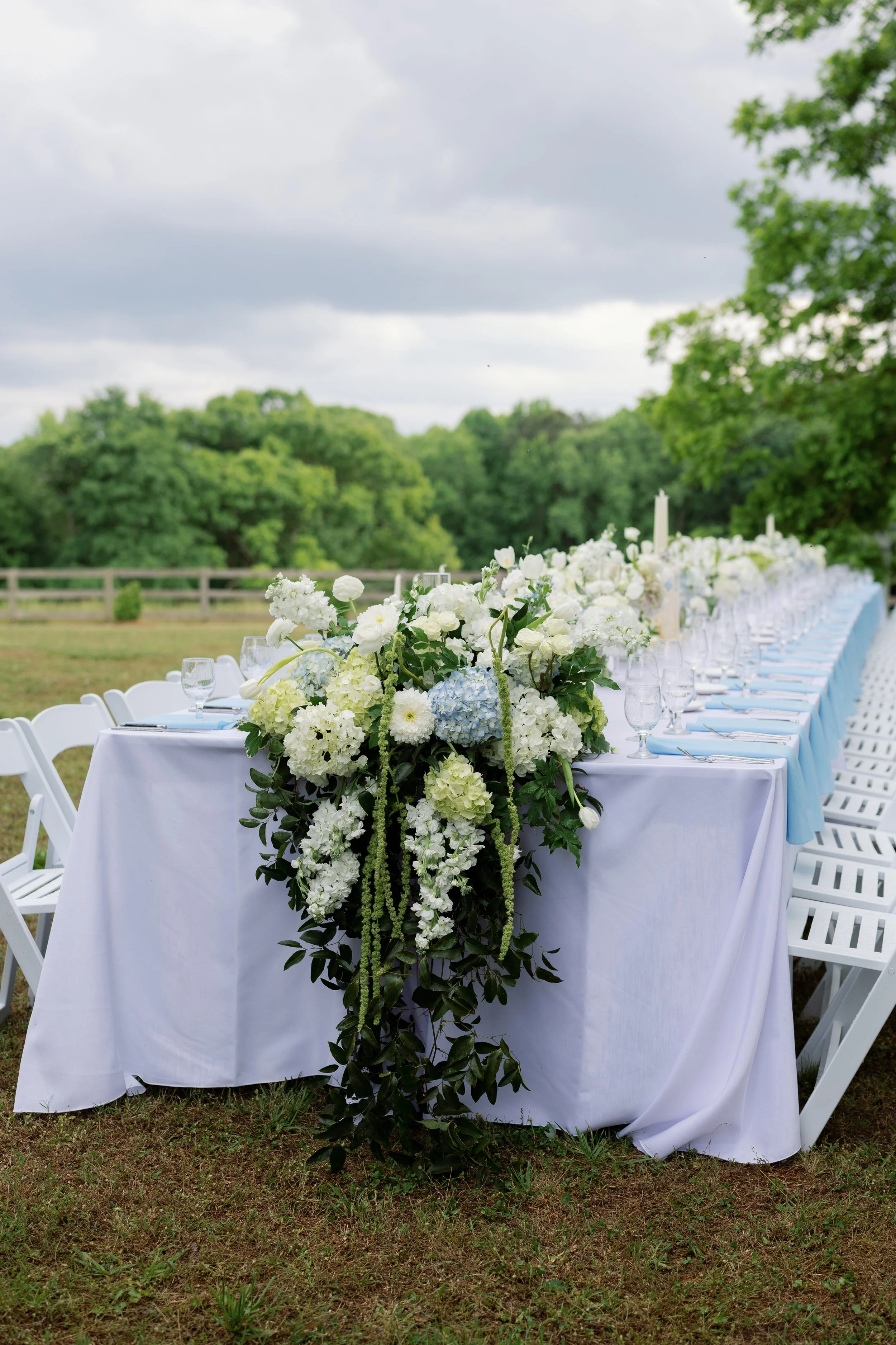Outdoor wedding reception table with white floral centerpiece, place settings, and white chairs in a garden