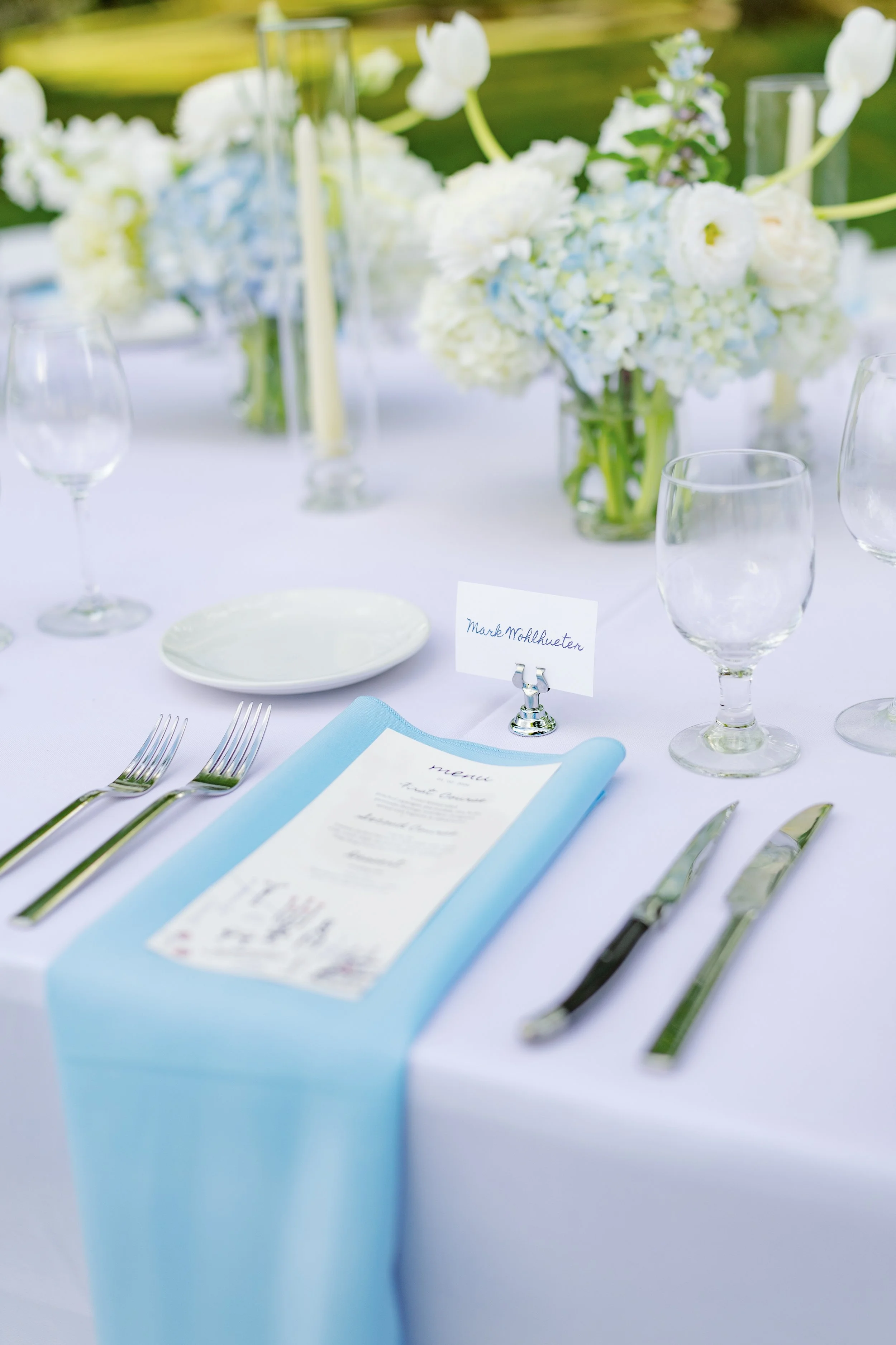 Elegant wedding reception table set with white and blue flowers, glassware, silverware, place settings, and a handwritten name card.