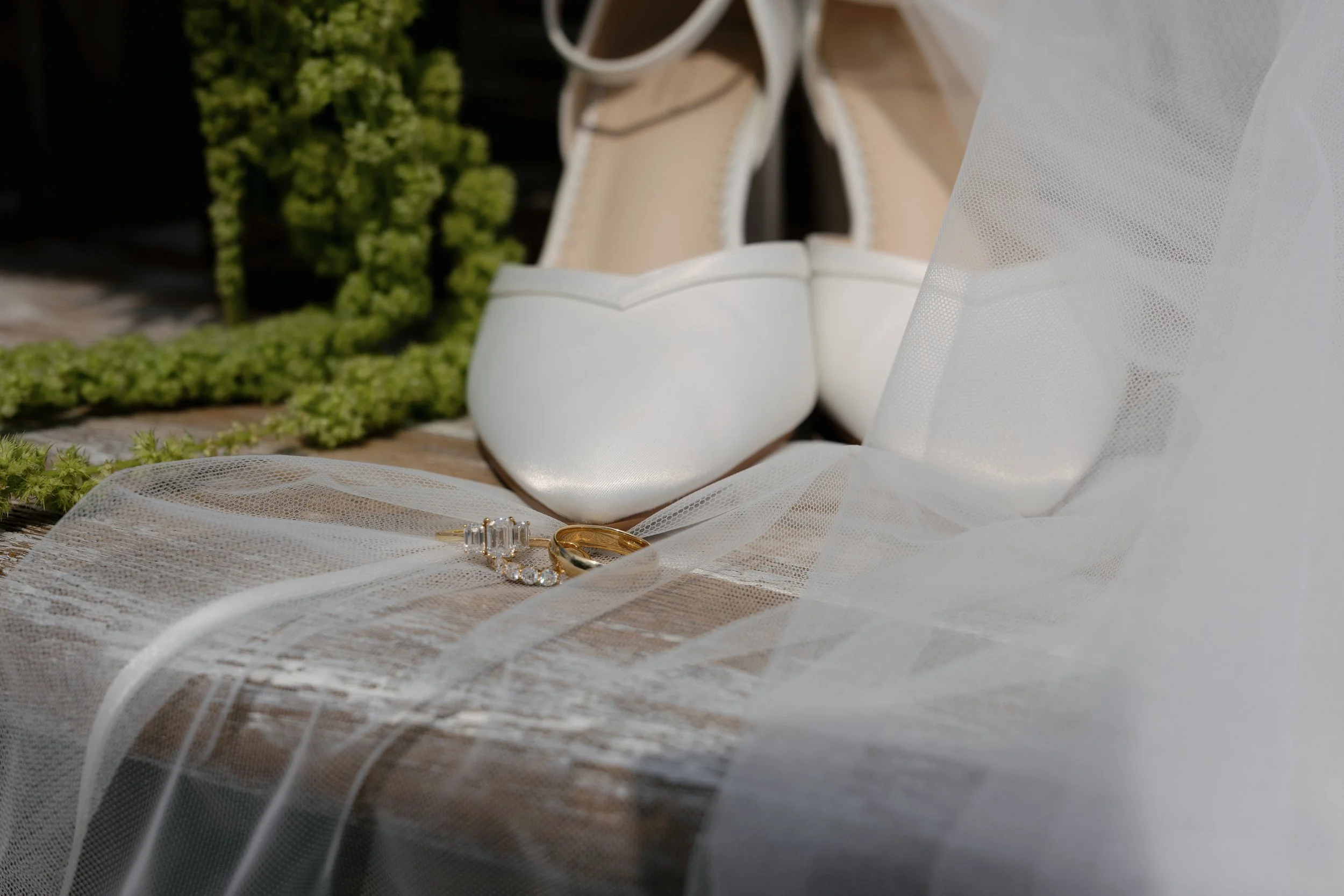 Close-up of white bridal heels, wedding rings, and sheer veil on a rustic wooden surface