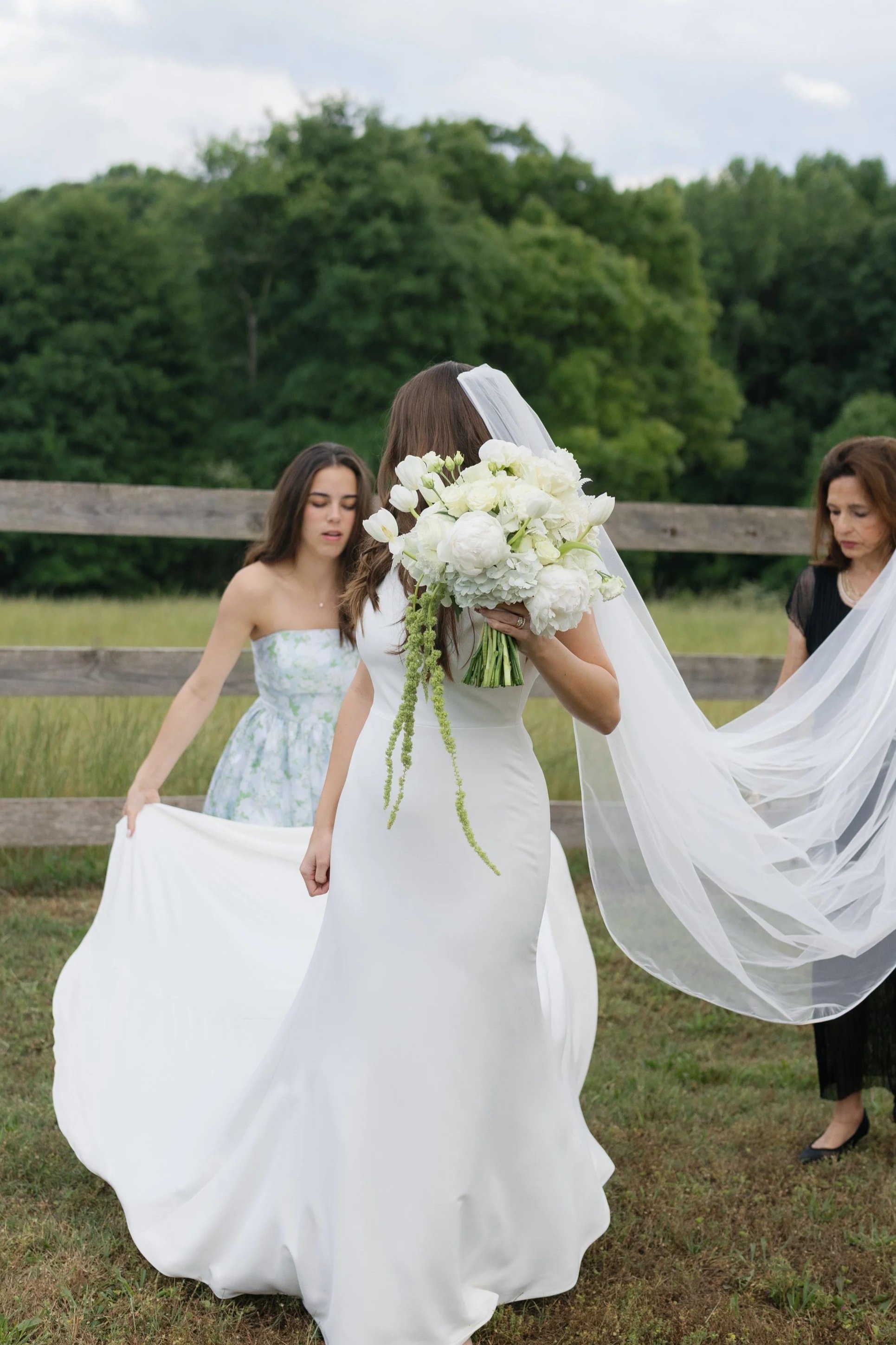 Bride in a white wedding dress and veil holding a large white bouquet outdoors, with attendants arranging her train.