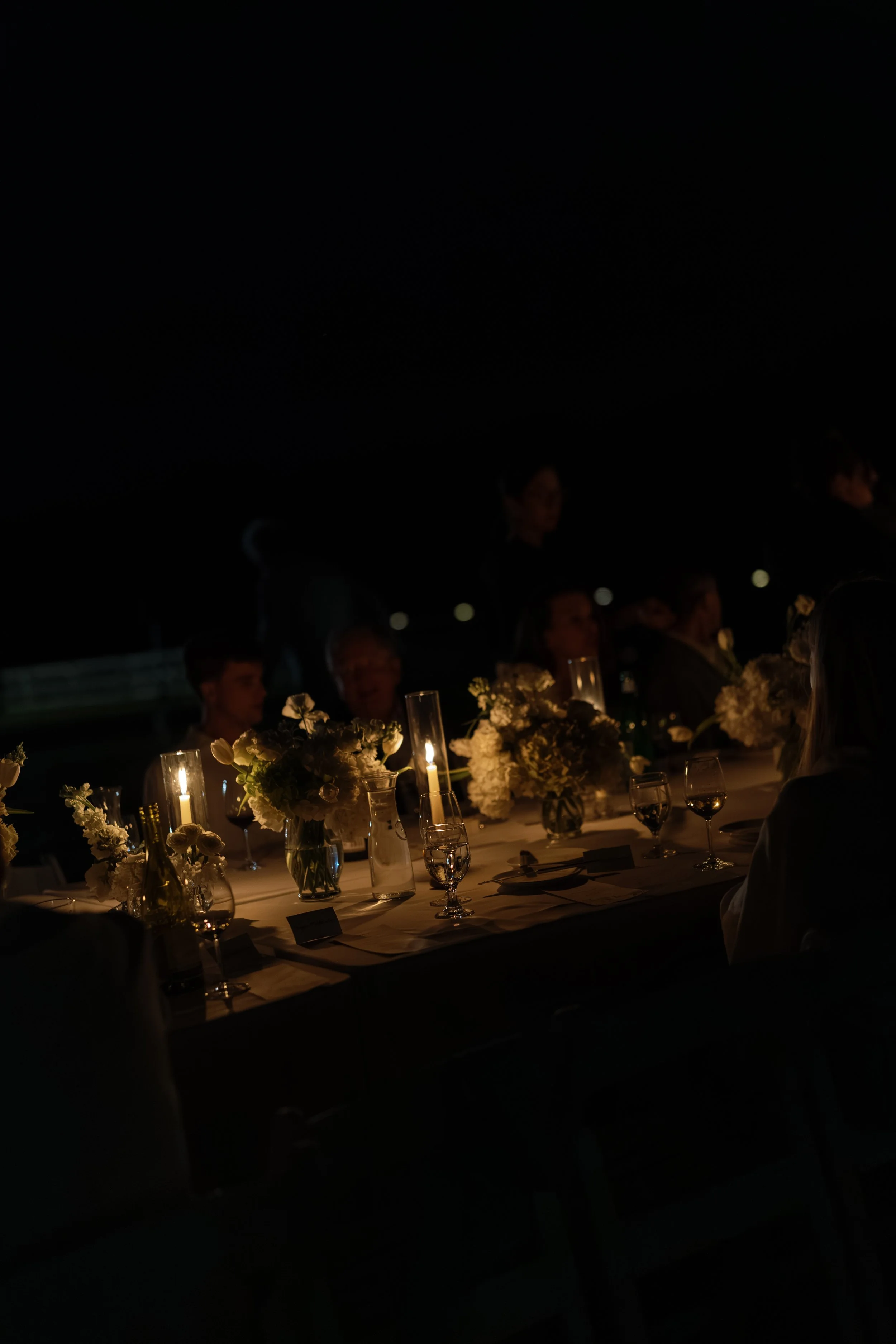 Dimly lit dinner party table with candles, flowers, wine glasses, and seated guests