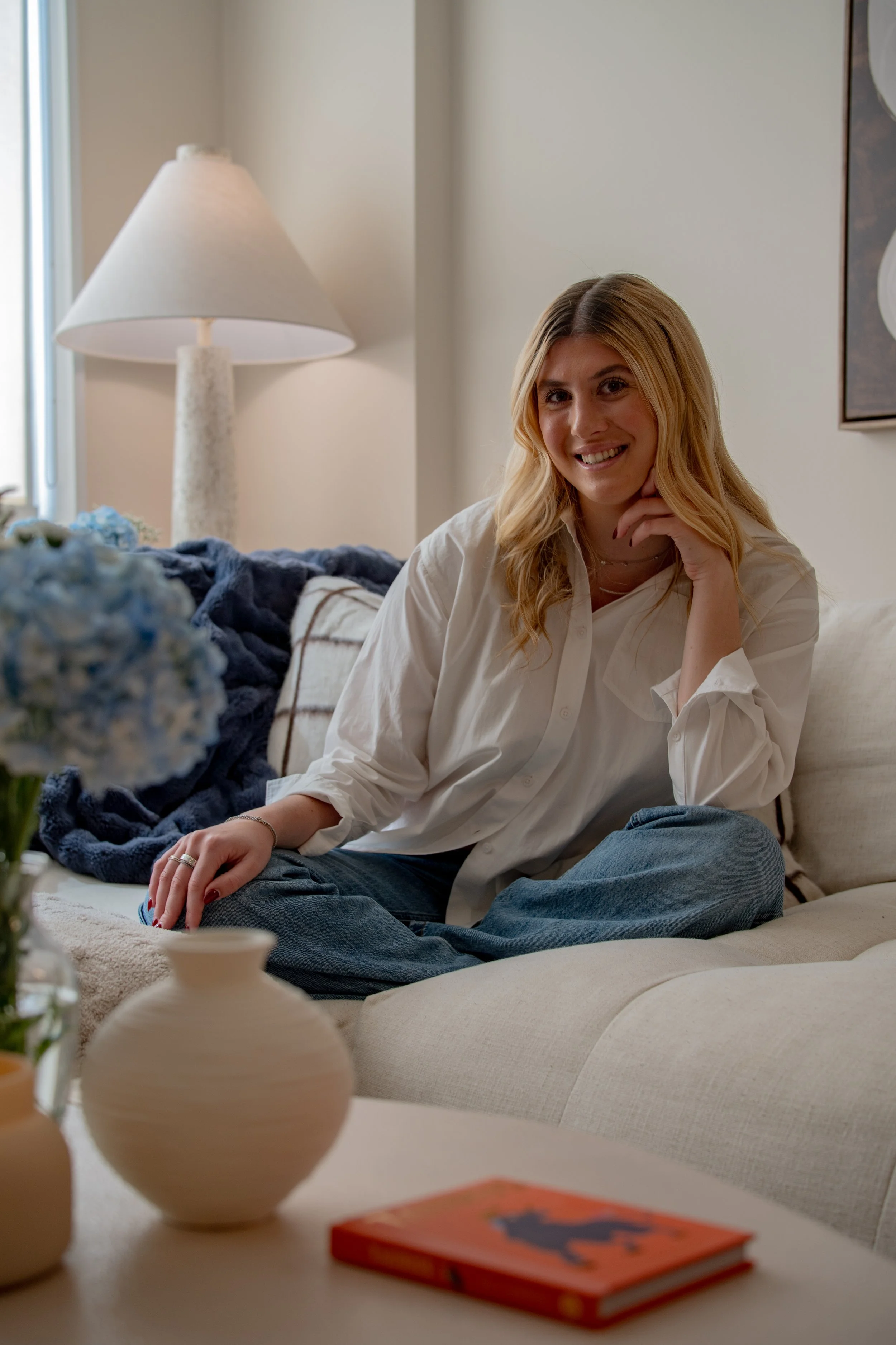 A woman with blonde hair sitting on a beige couch, smiling, in a cozy living room with a white table, vases, a lamp, and a decorative wall art.