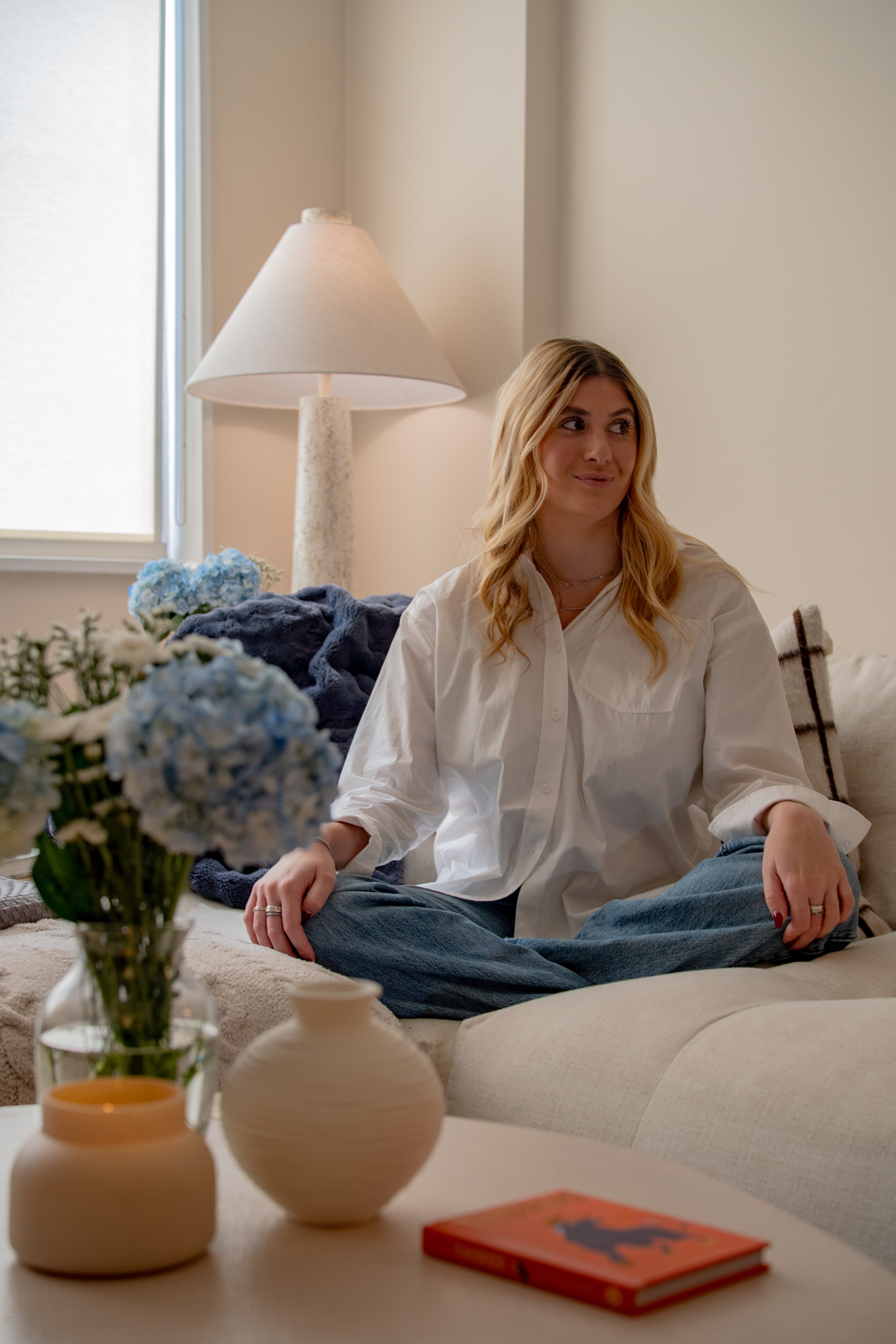 A woman with blonde hair sitting cross-legged on a beige sofa, wearing a white shirt and blue jeans in a well-lit room with beige walls and a window. There are blue hydrangea flowers, a navy blanket, vases, and a book with an orange cover on a table 
