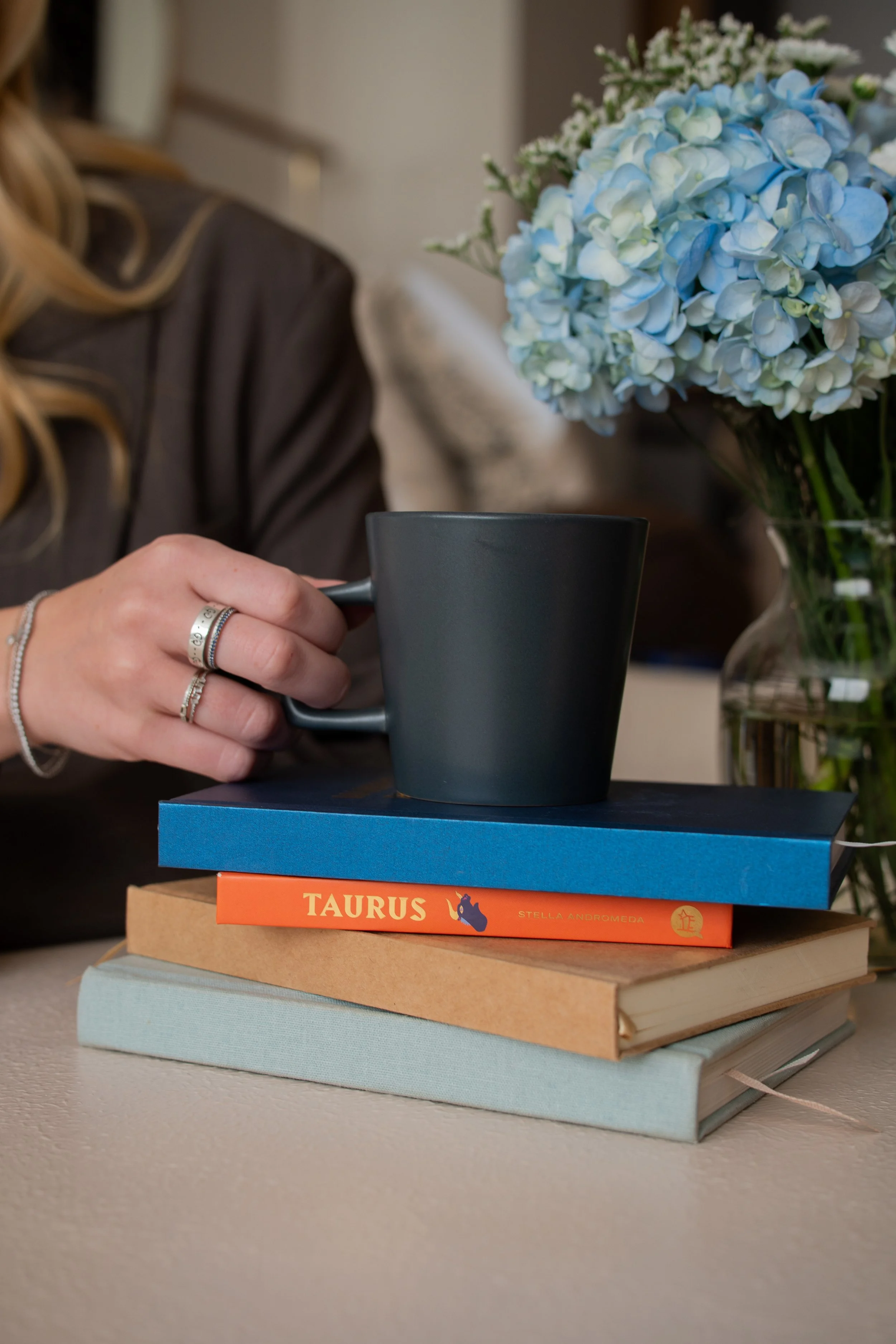 A person's hand holding a dark ceramic mug over a stack of three books on a beige table. The books are titled, from top to bottom: "Taurus" by Stella Andromeda, with an orange cover; a blue-covered book; and a large light-colored hardcover. In the ba