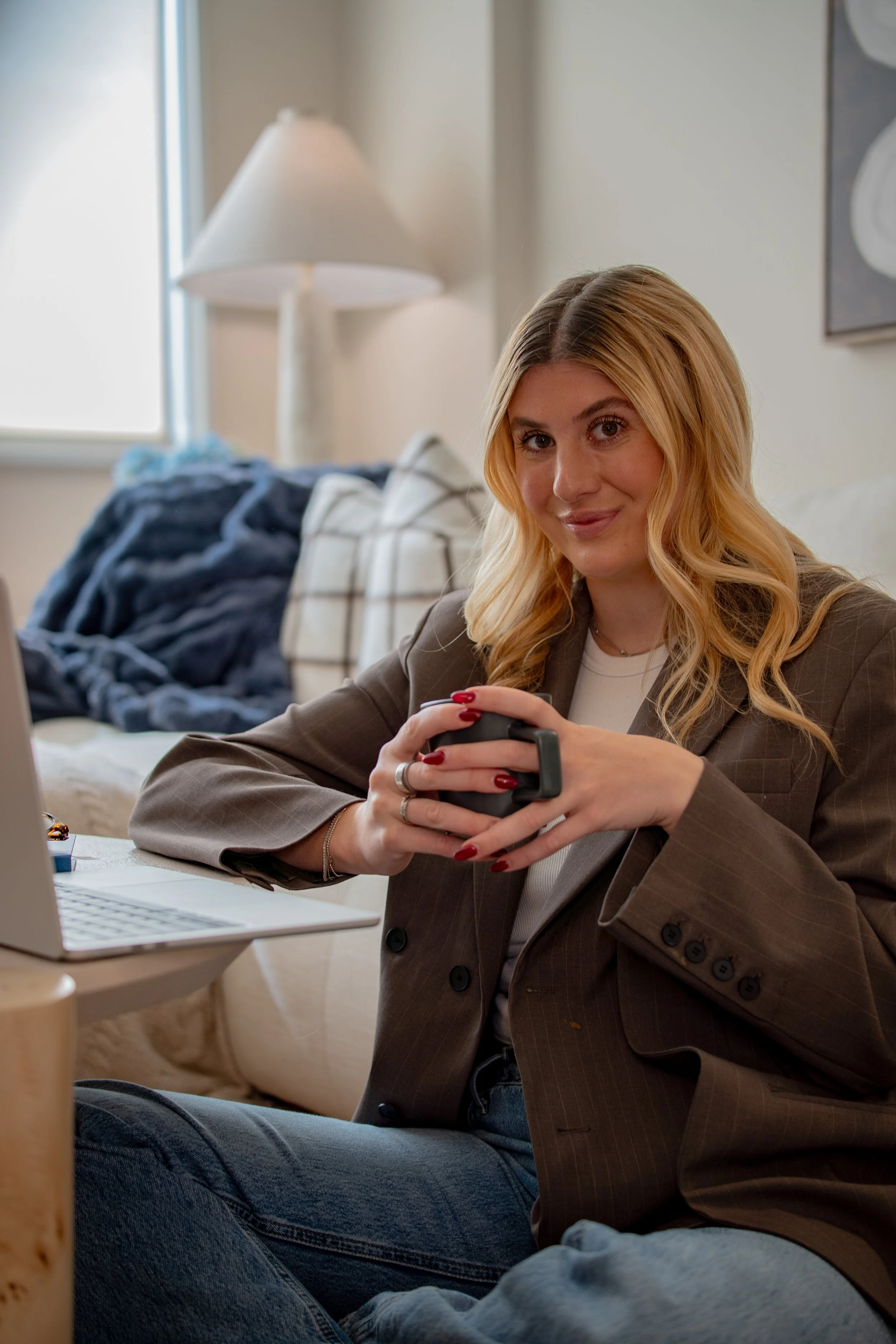 A woman with blonde hair, wearing a brown blazer and white shirt, sitting on a couch and smiling while holding a smartphone in her hands. There are cushions and a blanket on the couch, a lamp and wall art in the background, and a laptop on the table in front of her.