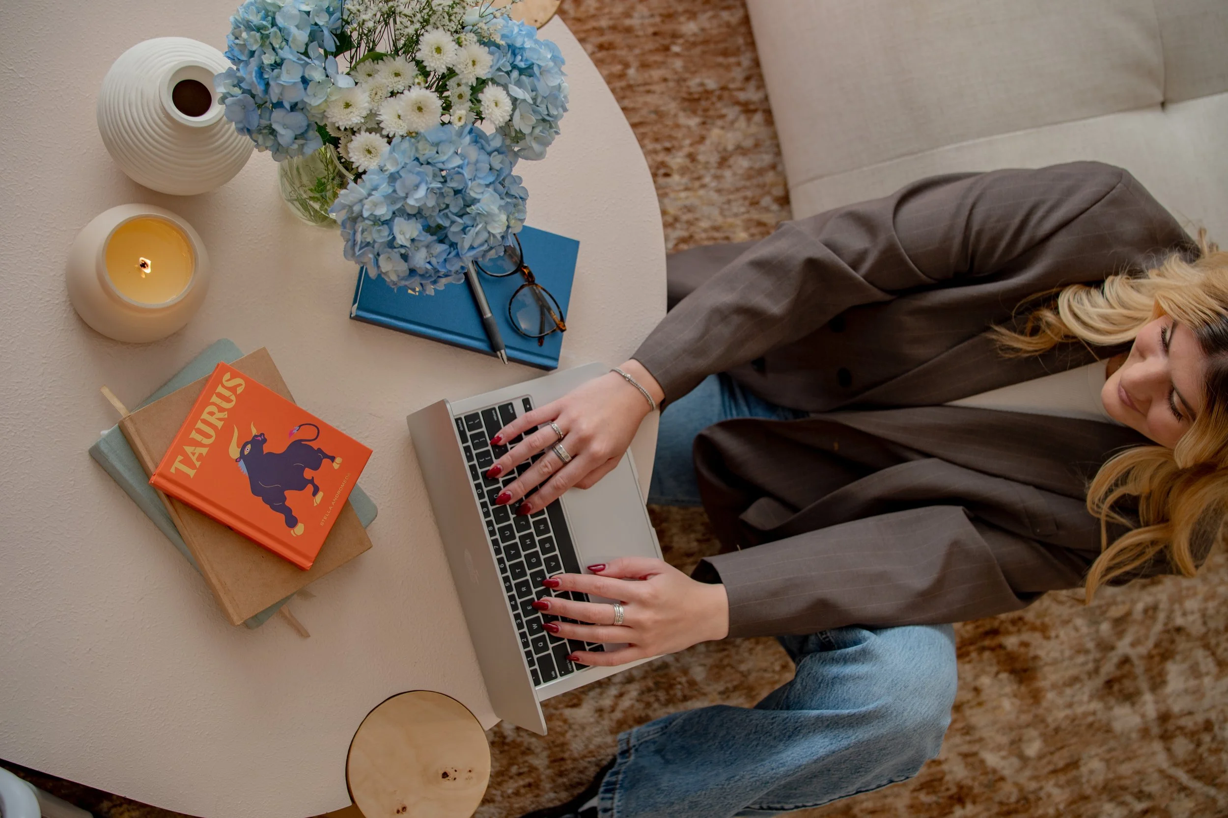 A woman with blonde hair wearing a gray blazer and blue jeans sitting at a round white table, using a silver laptop. The table has a vase of blue and white flowers, a white ribbed vase, a lit candle, two books (one titled 'TAURUS' with a black bull illustration), a pair of eyeglasses, a blue notebook, and a desk pen.