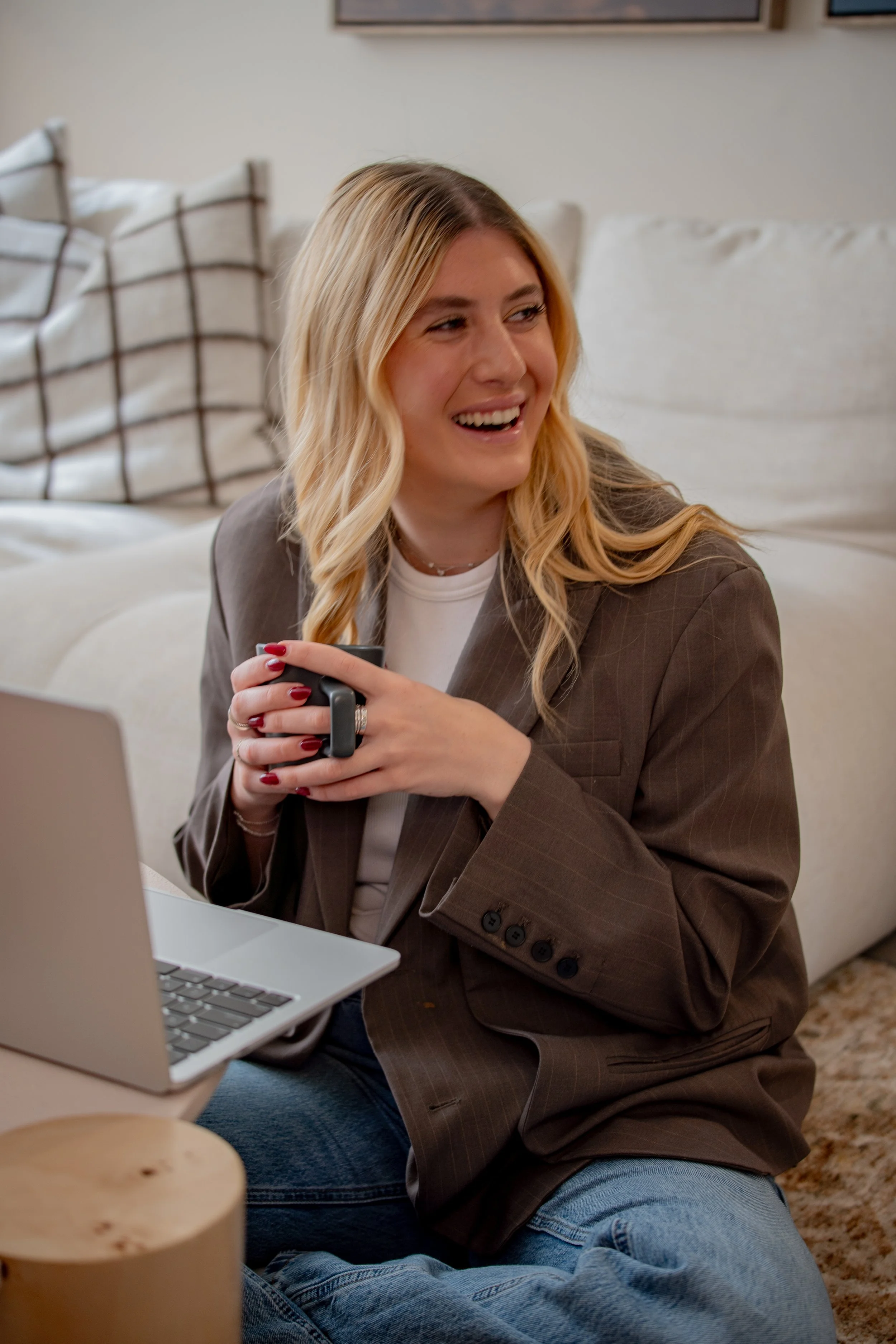 A woman with blonde hair and a brown blazer sitting on a couch, holding a mug, smiling, with a laptop on her lap.